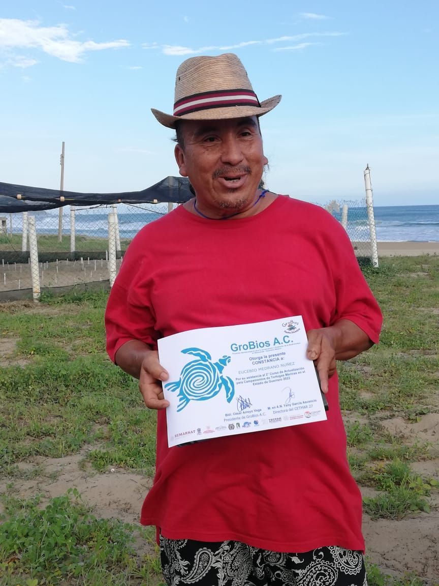 Man in a red shirt and straw hat holding a certificate outdoors by the water