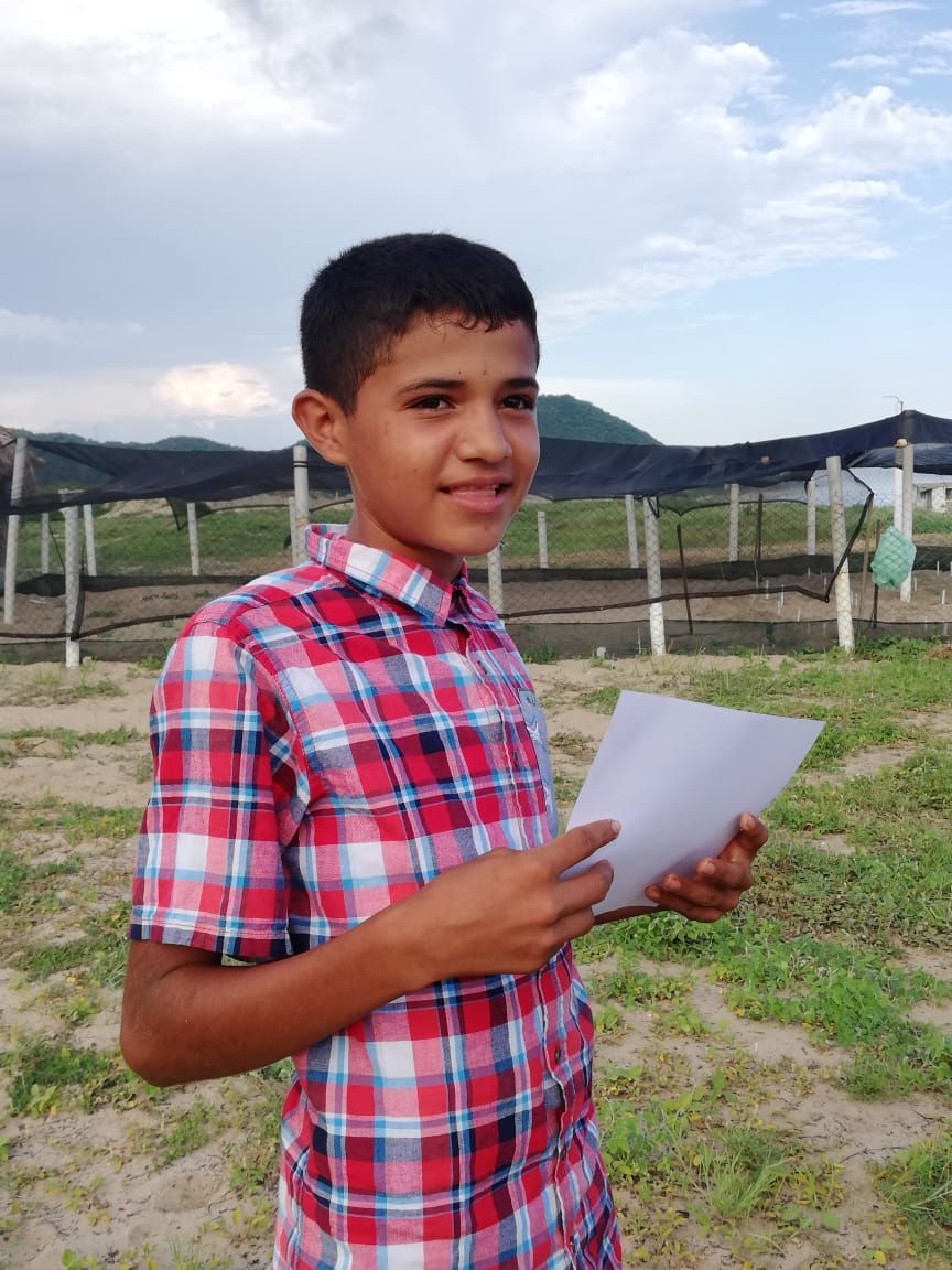 Young person in a red plaid shirt holding a white paper outdoors near hills and fences