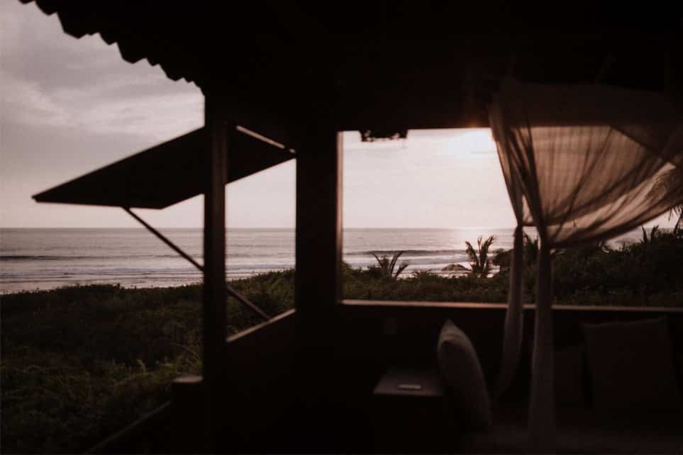 Dim seaside veranda at sunset, with silhouetted posts, curtains, and ocean horizon.