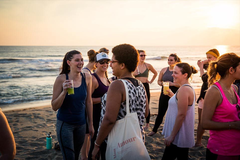 People socializing on a beach at sunset, holding drinks and chatting near the shoreline.