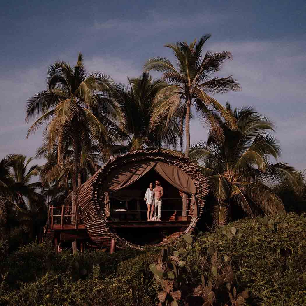 Two people standing on a rustic hut deck amid palm trees at dusk
