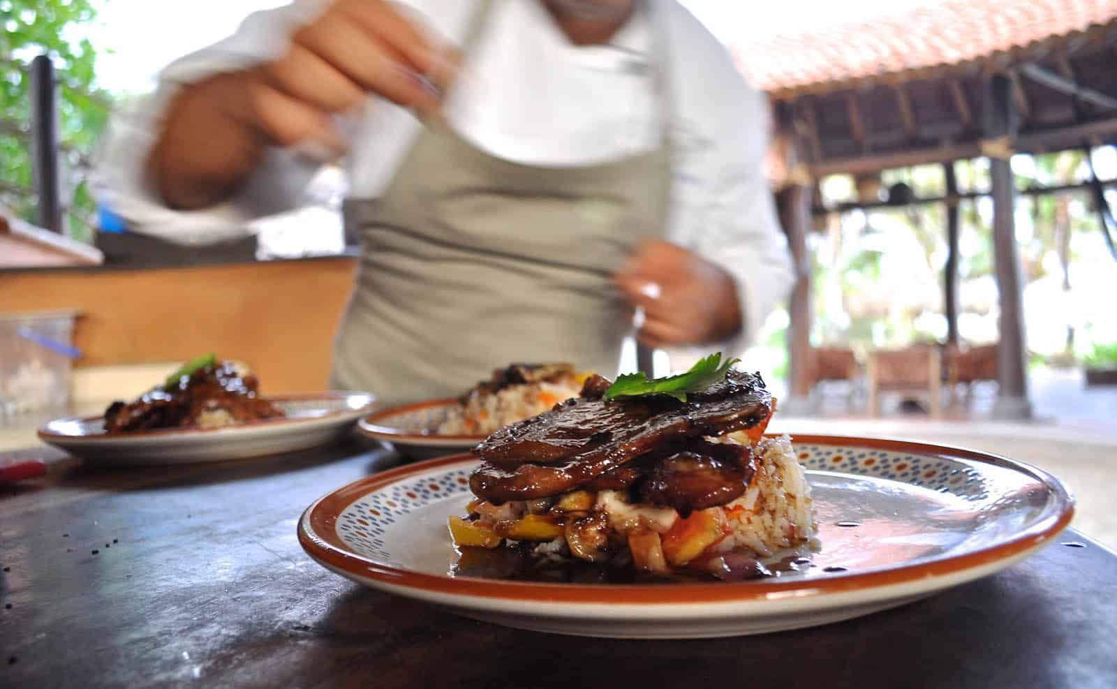 Chef plating a dish on a restaurant table, with two finished meals in the foreground.