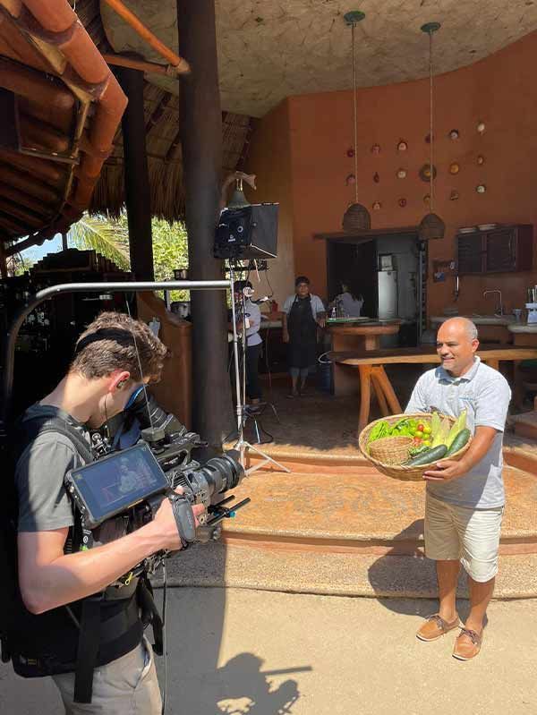 Cameraman filming a man holding a green basket in a rustic open-air setting