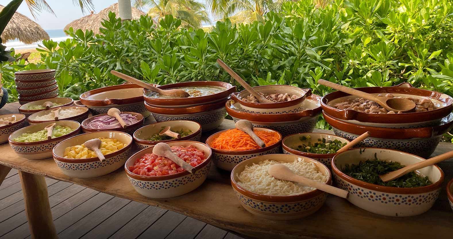 Assorted colorful spices and ingredients in woven bowls on a table outdoors, with tropical greenery behind.