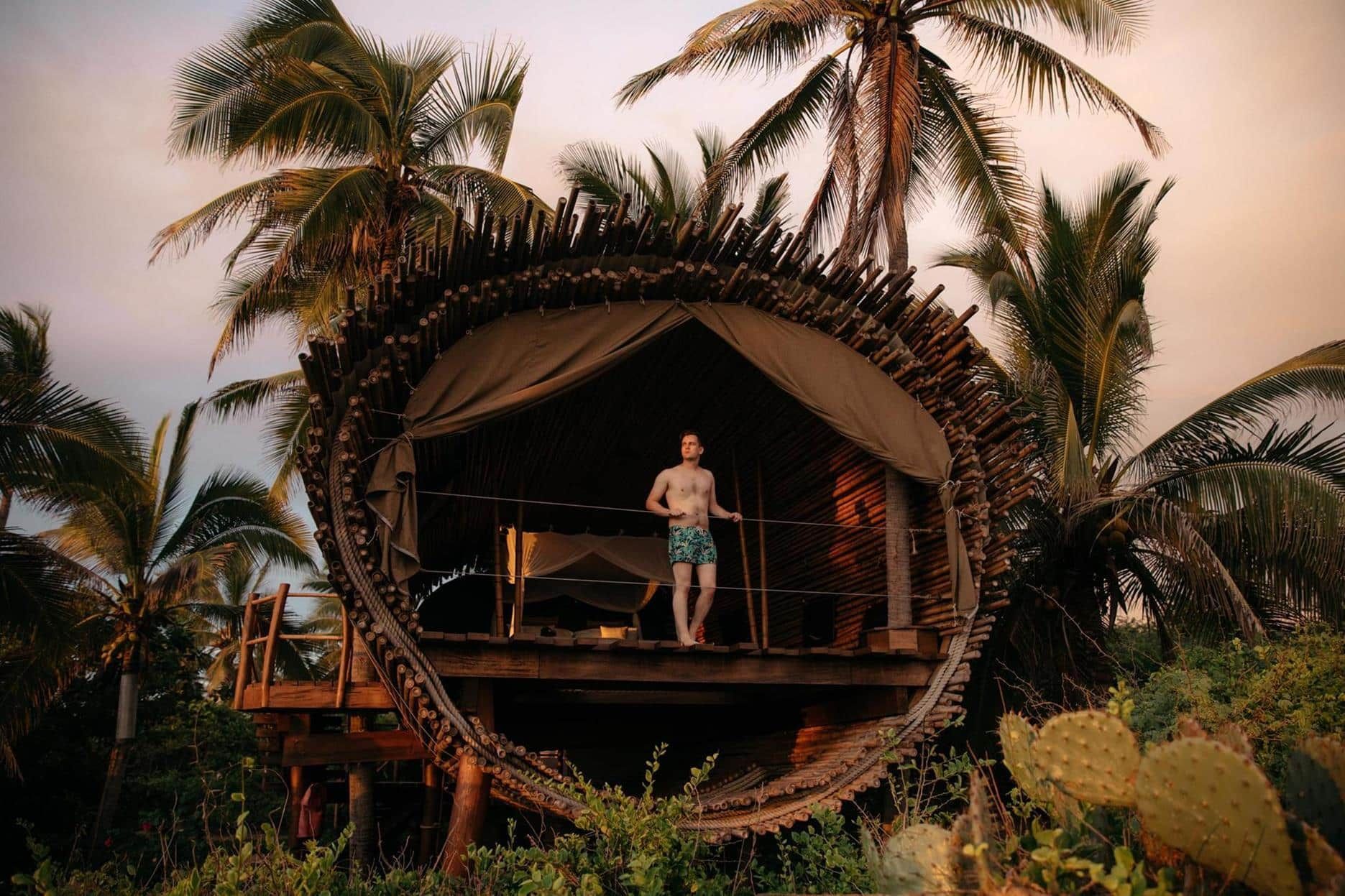 Person standing on a tropical hut deck surrounded by palm trees at sunset