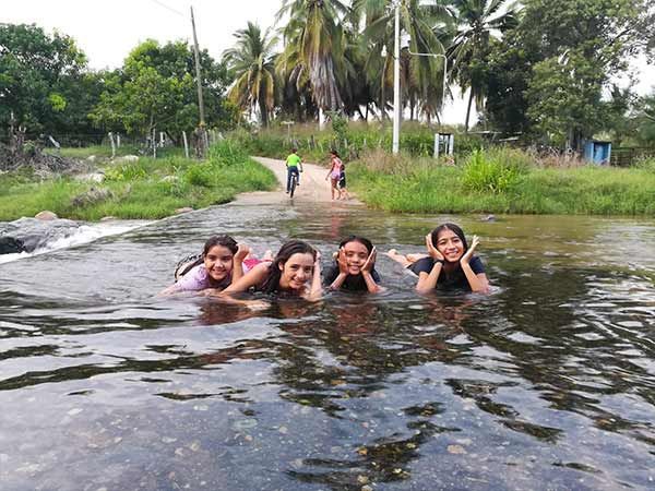 Four people smiling in a river, floating in water with lush green trees and a rural path in the background.