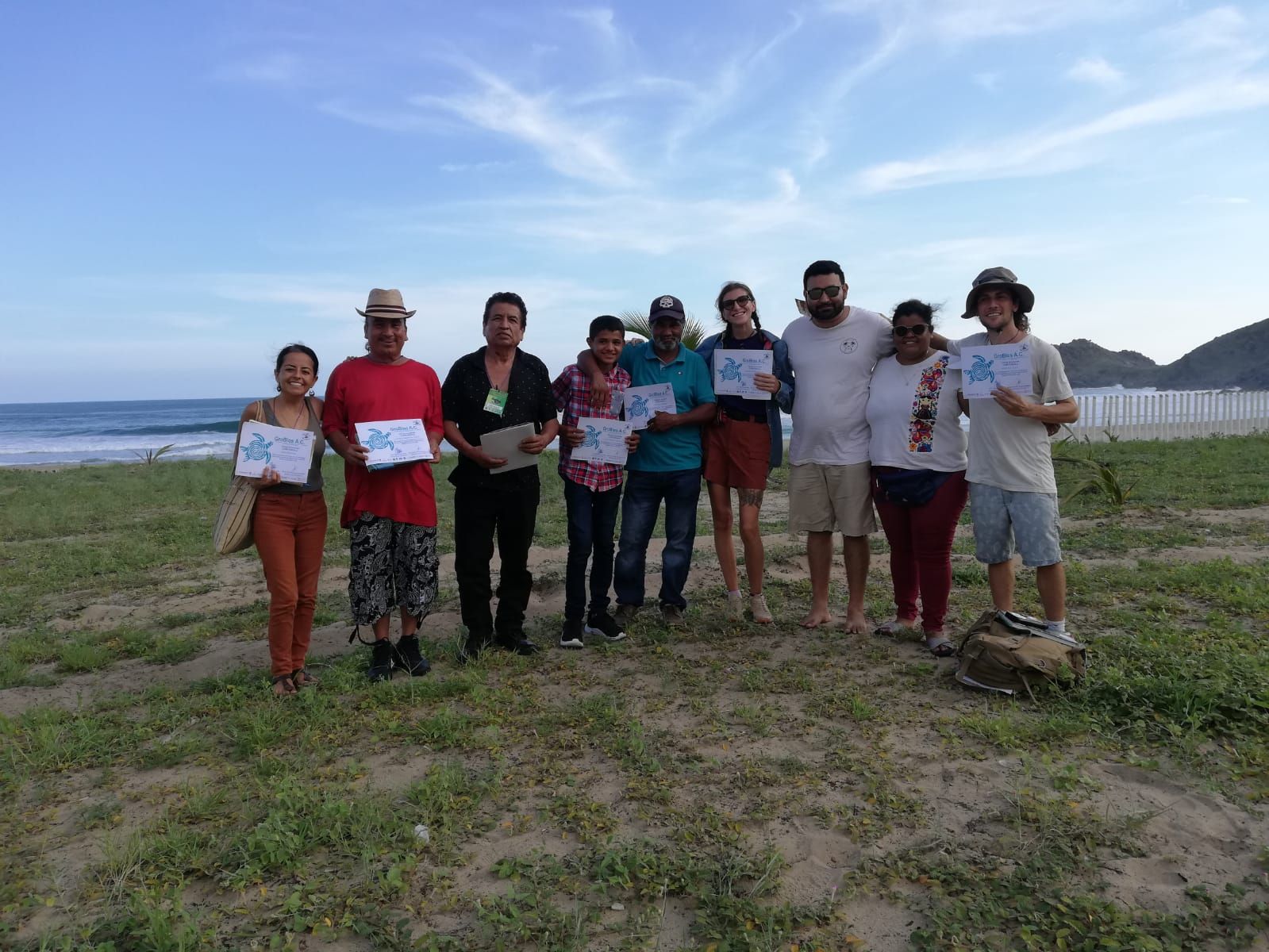 Group of people standing outdoors on grass holding certificates, with water and hills in the background.