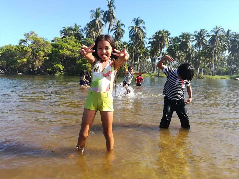Children playing in shallow river water with palm trees and greenery in the background