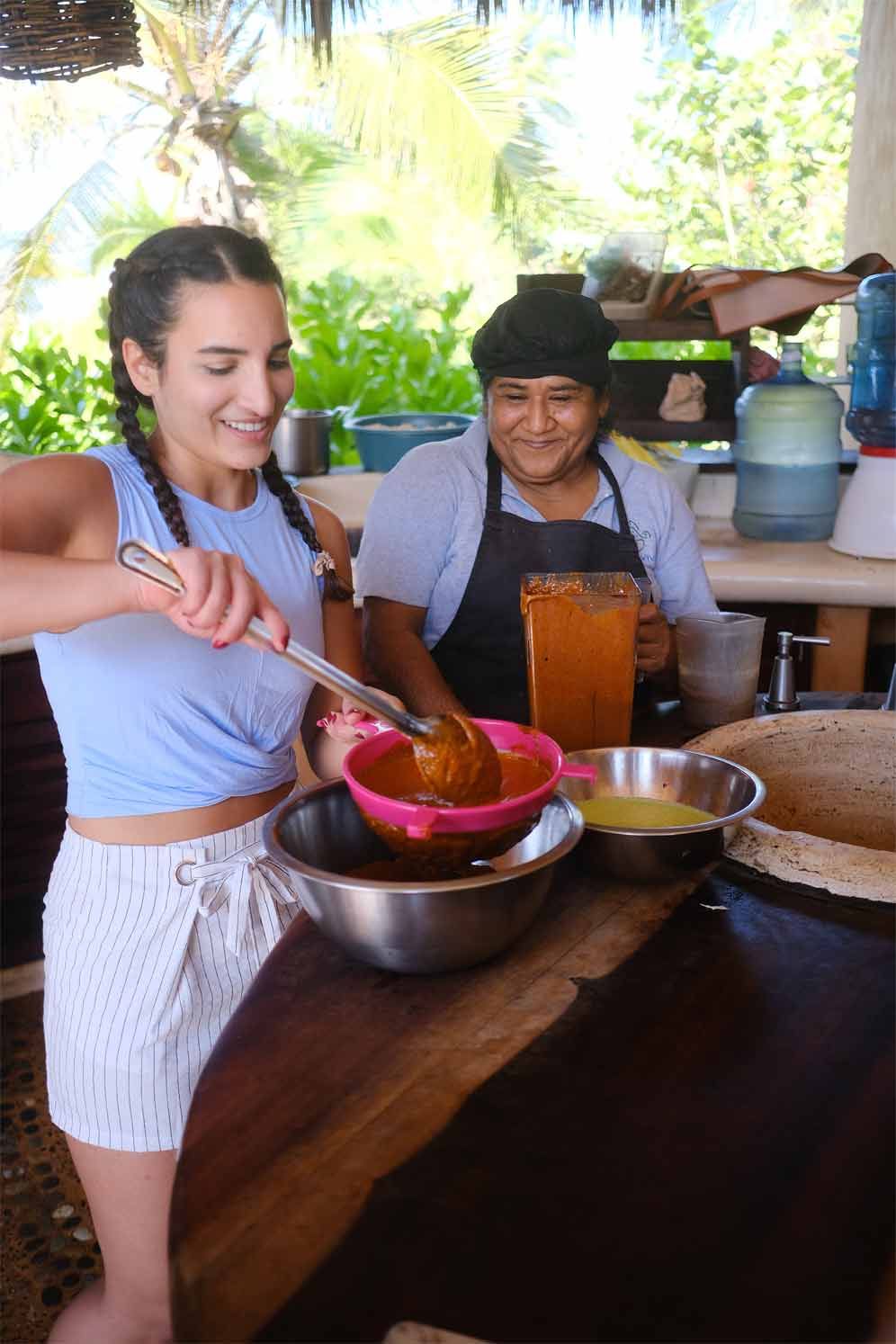 Two women preparing food at a wooden table, one stirring a bowl with a pink spoon in a bright open-air kitchen.
