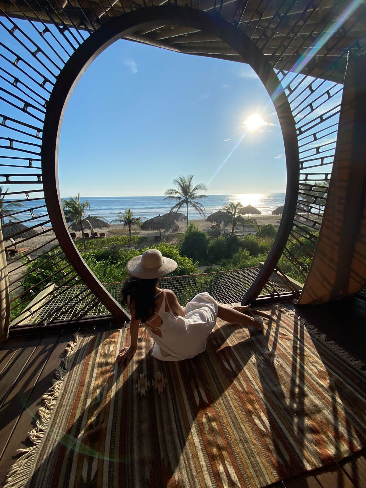 Woman in a sun hat sits in a circular window frame, overlooking a tropical beach and ocean at sunset.