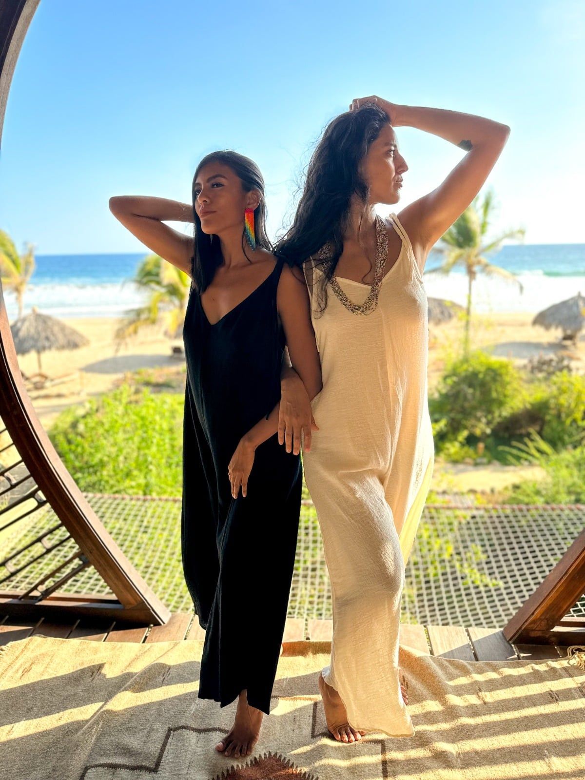Two women posing on a beachside deck, one in black and one in cream, with ocean and palm trees behind them