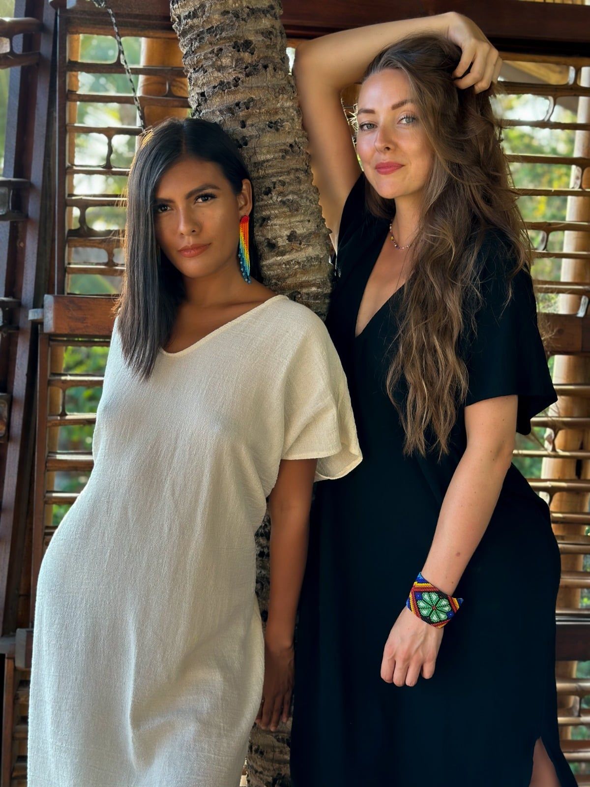 Two women posing by a tree indoors, one in a cream dress and one in a black dress.