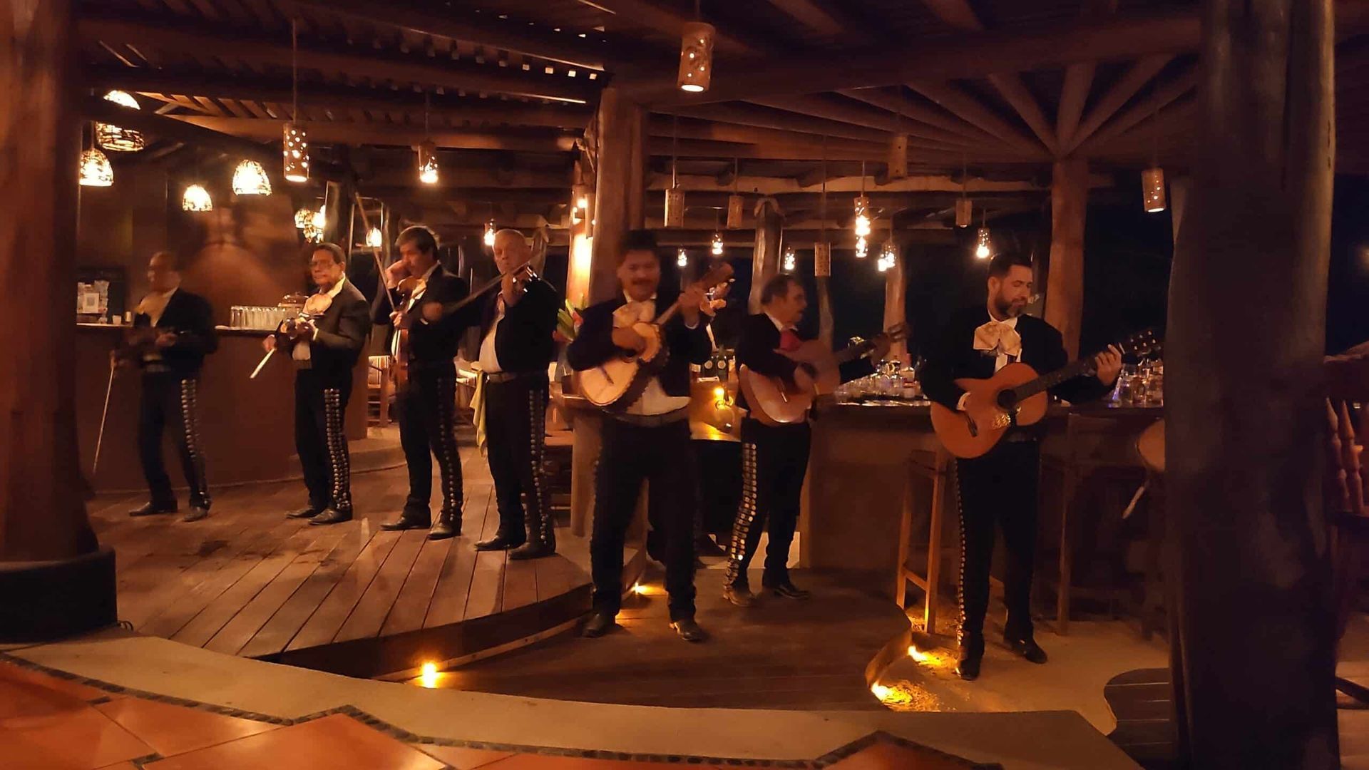 Live band performing on a warmly lit wooden stage in a rustic hall, with guitars and drums.