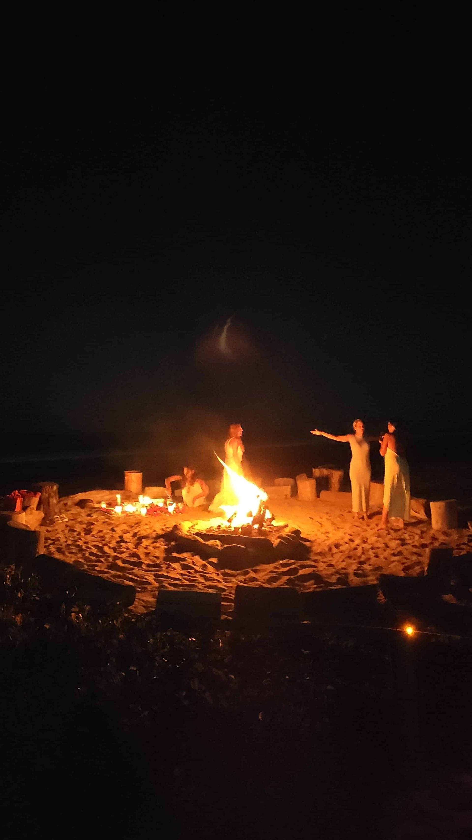 People gathered around a bright campfire at night in a dark outdoor setting.