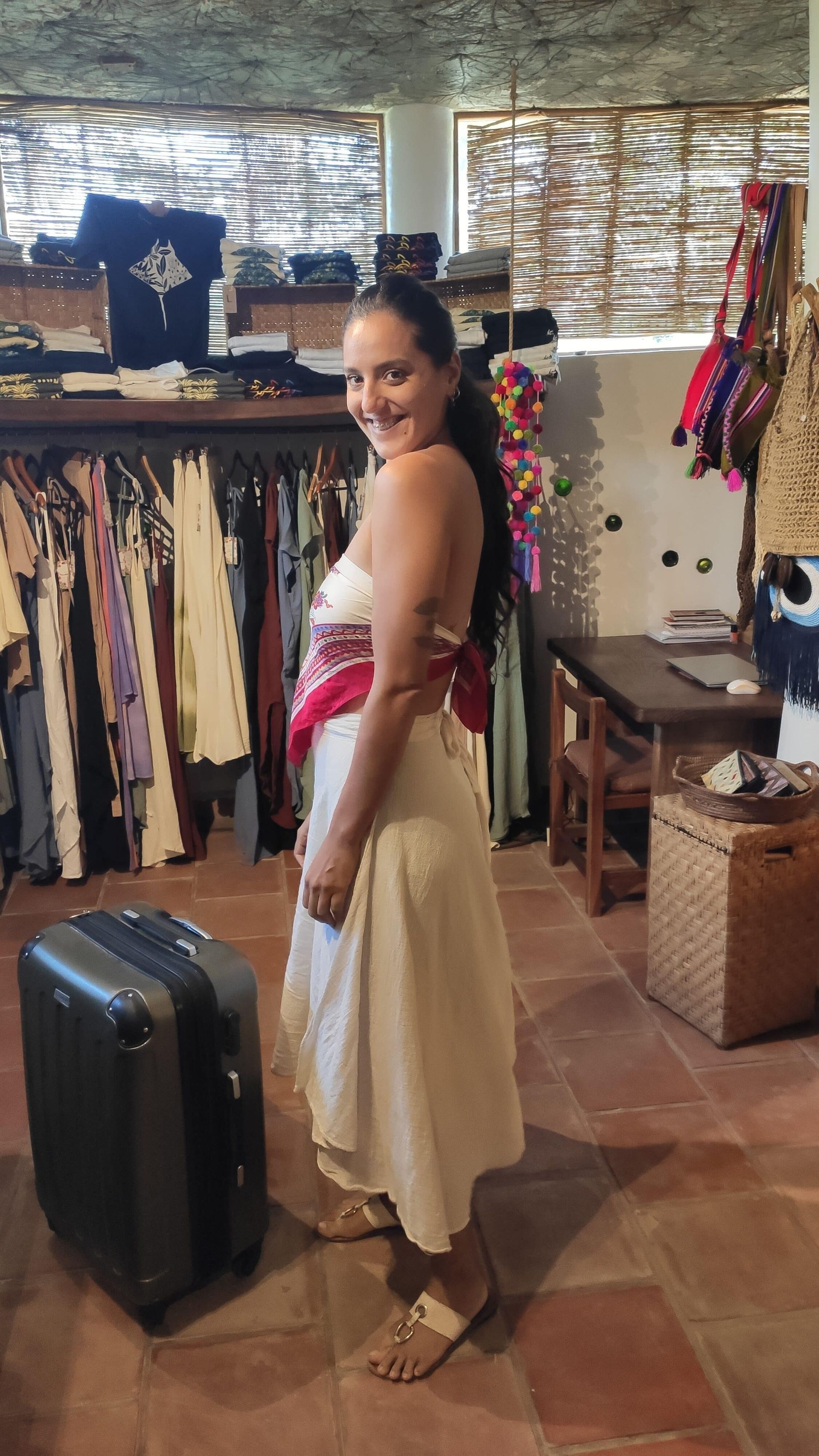 Woman posing in a boutique, wearing a cream dress with a pink sash beside a black suitcase.