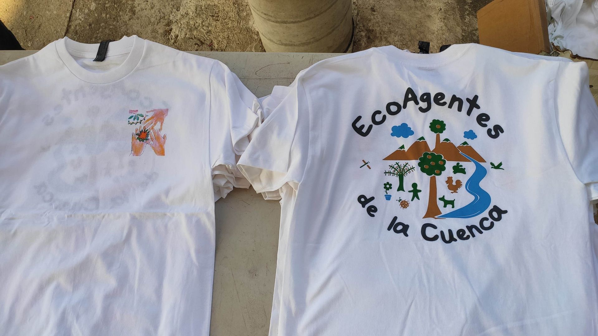 Two white T-shirts laid side by side; one reads “EcoAgentes de la Cuenca” with a tree and water graphic.