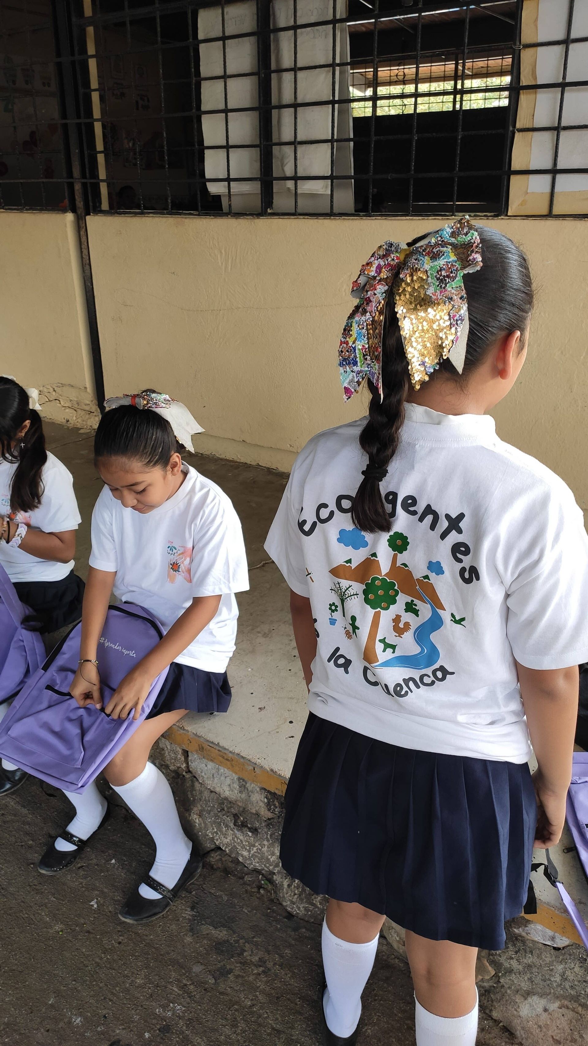 Students in white uniforms sit and stand beside a school bench in a hallway.