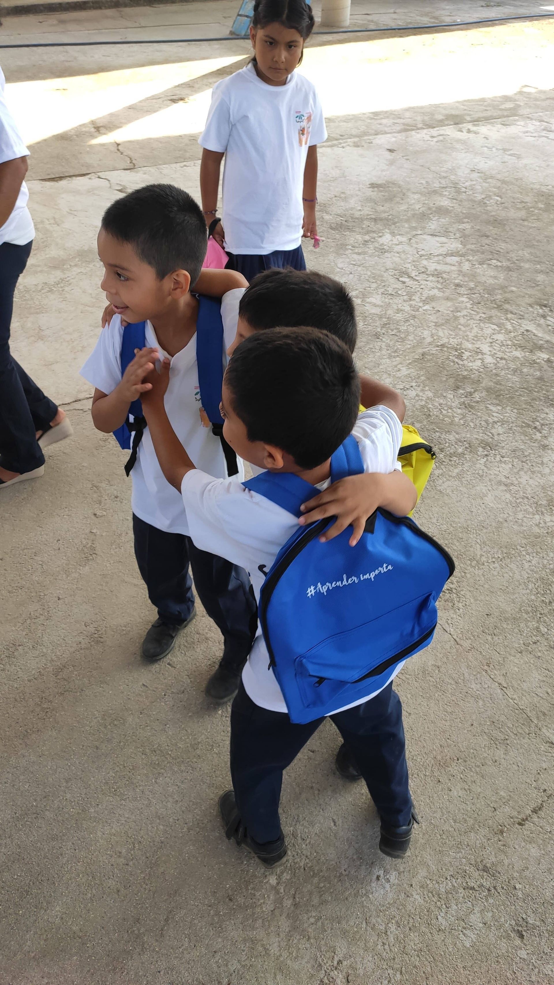 Three schoolchildren in white shirts and navy pants walk together outdoors, one with a blue backpack.