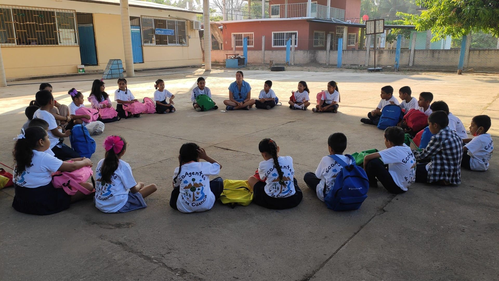 Children sitting in a large circle outdoors in a school courtyard, facing inward for a group activity.