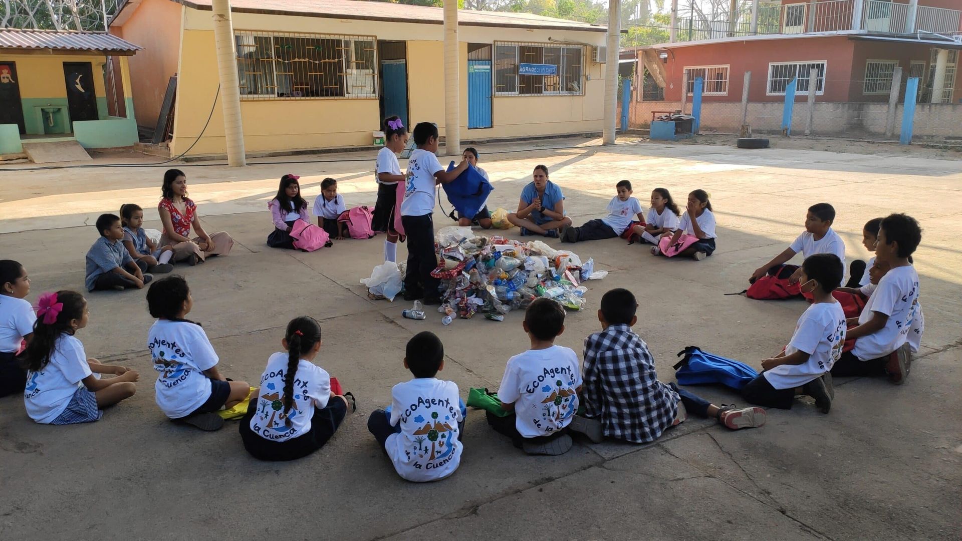 Children seated in a circle outdoors with a teacher at center, in a school courtyard
