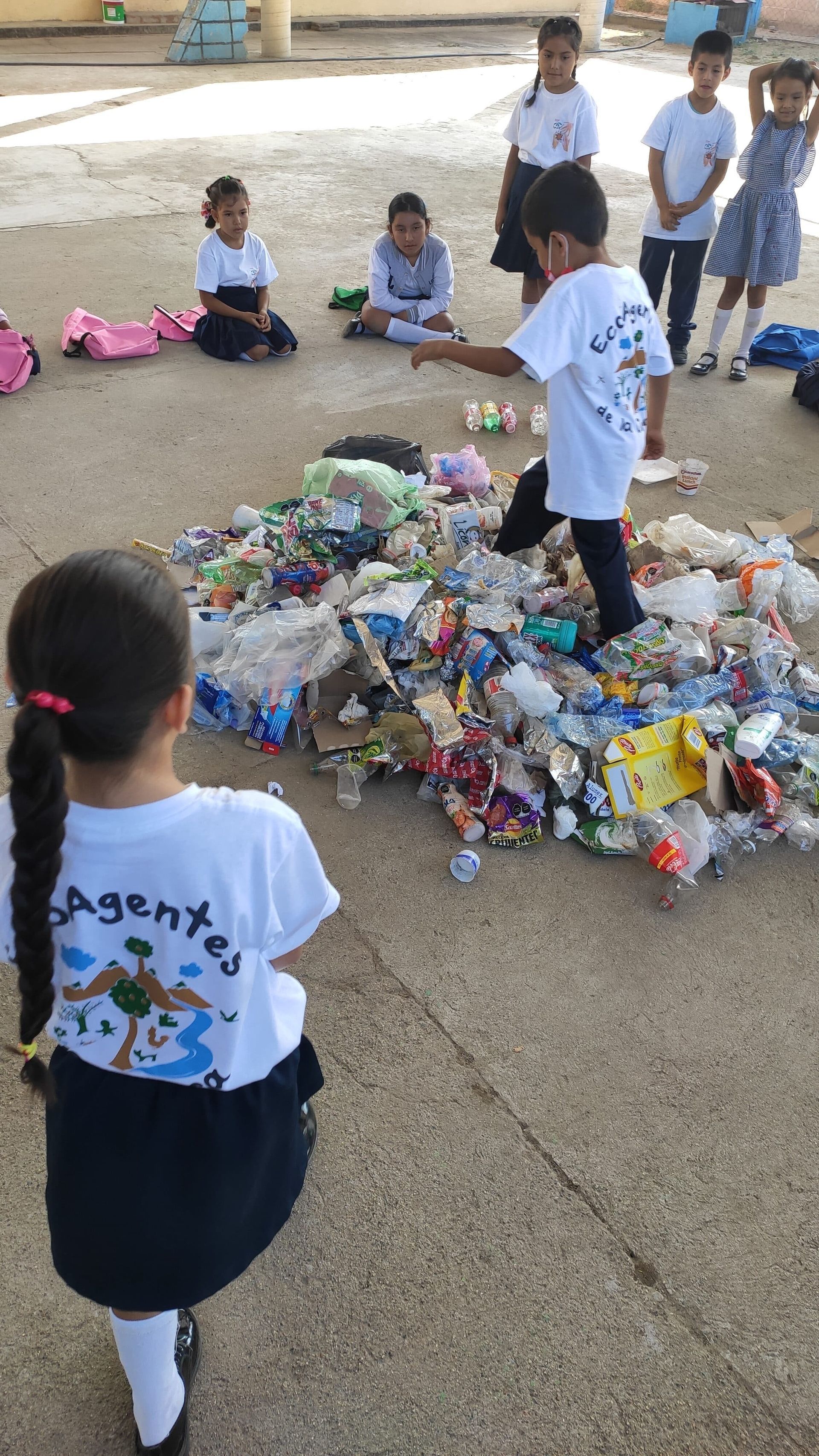 Children in school uniforms gather around a pile of plastic bottles for recycling