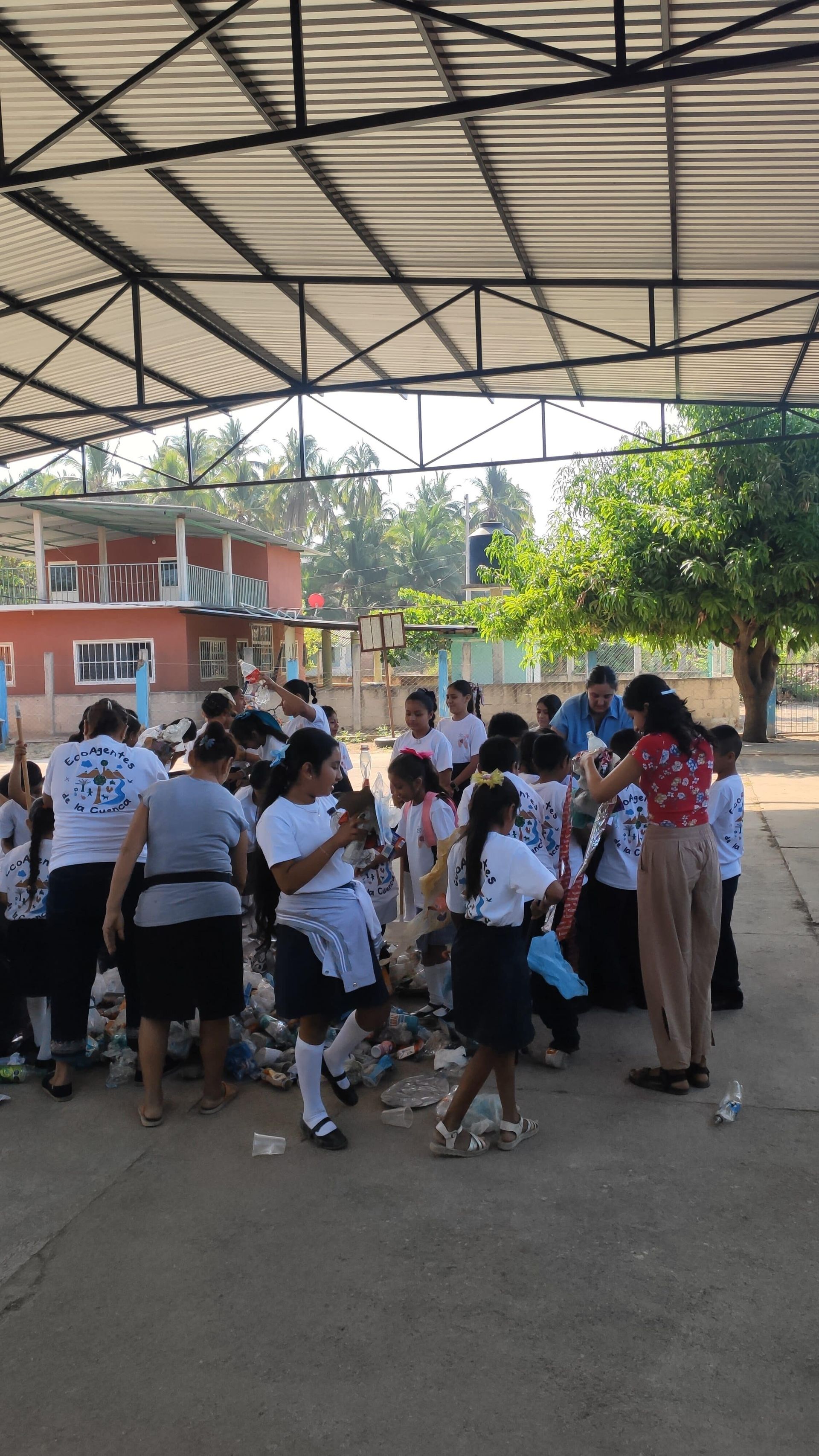 Group of people gathered outdoors under a metal roof, talking and moving near a red building and trees.