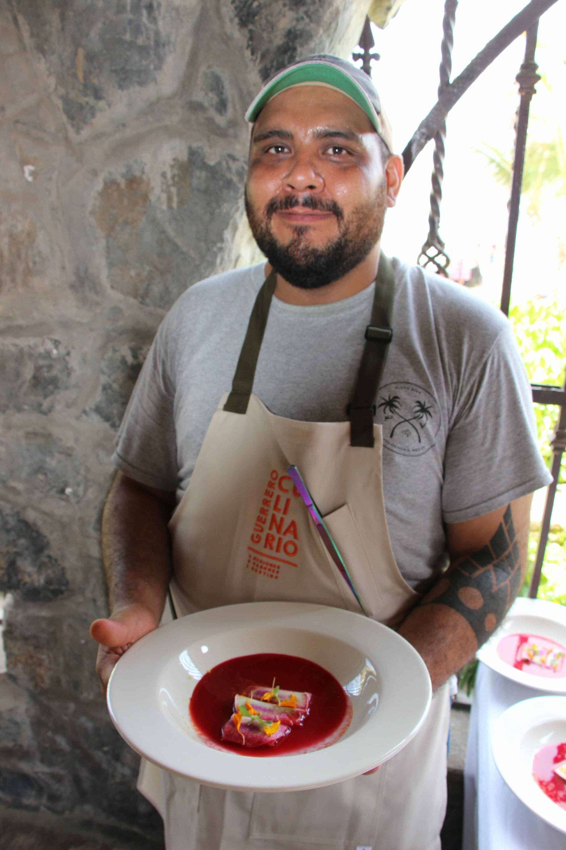 Chef in apron holding a plated dessert, standing indoors and smiling.