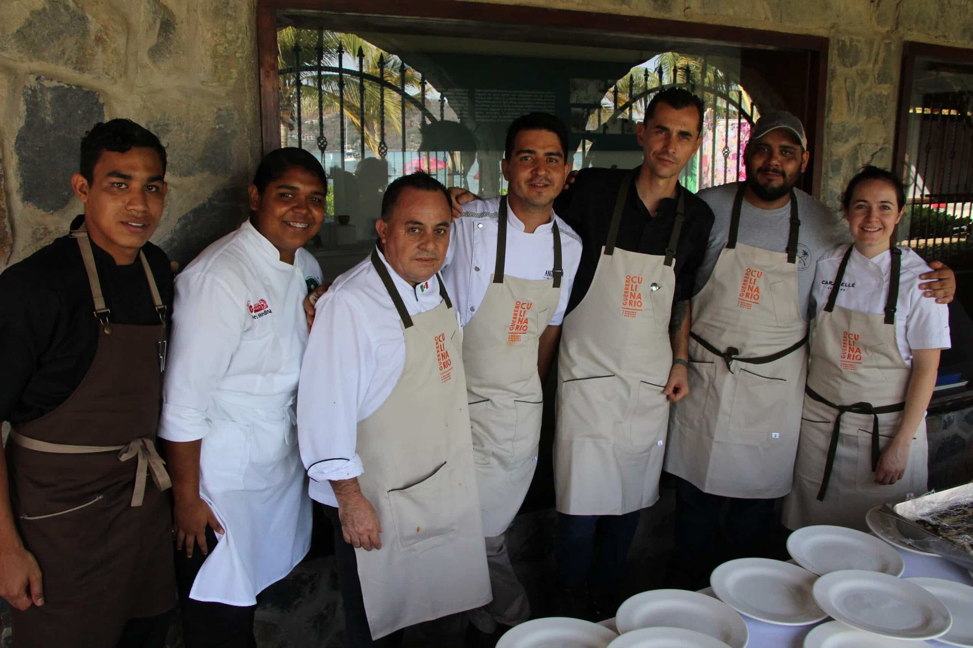 Seven restaurant staff in aprons and chef coats pose together in a kitchen with stacked plates.