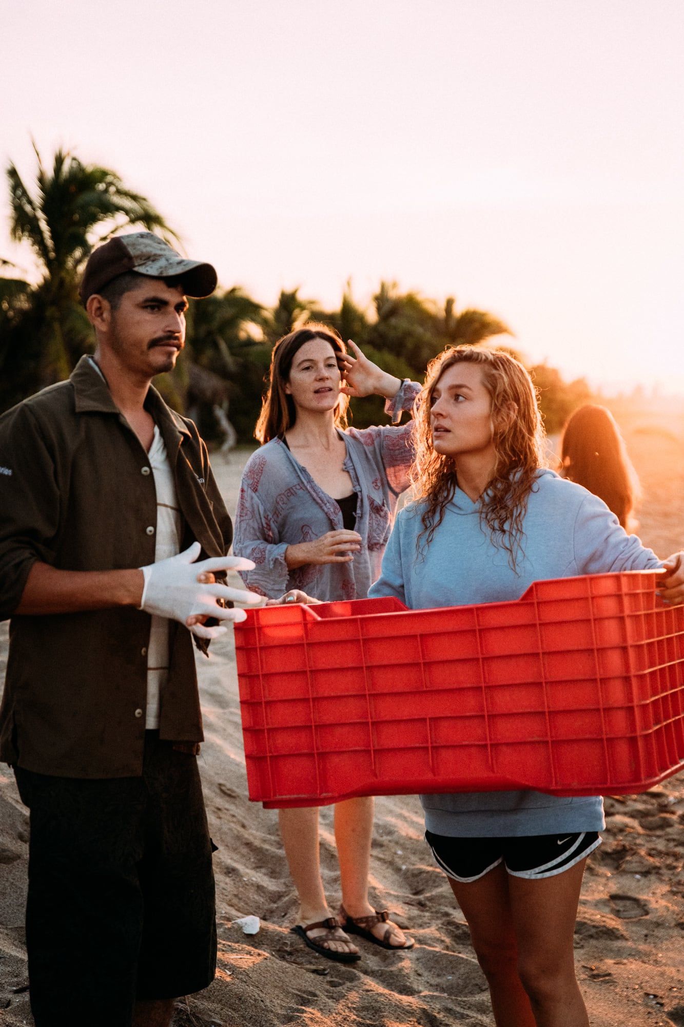 Three people carrying a red crate on a sandy beach at sunset