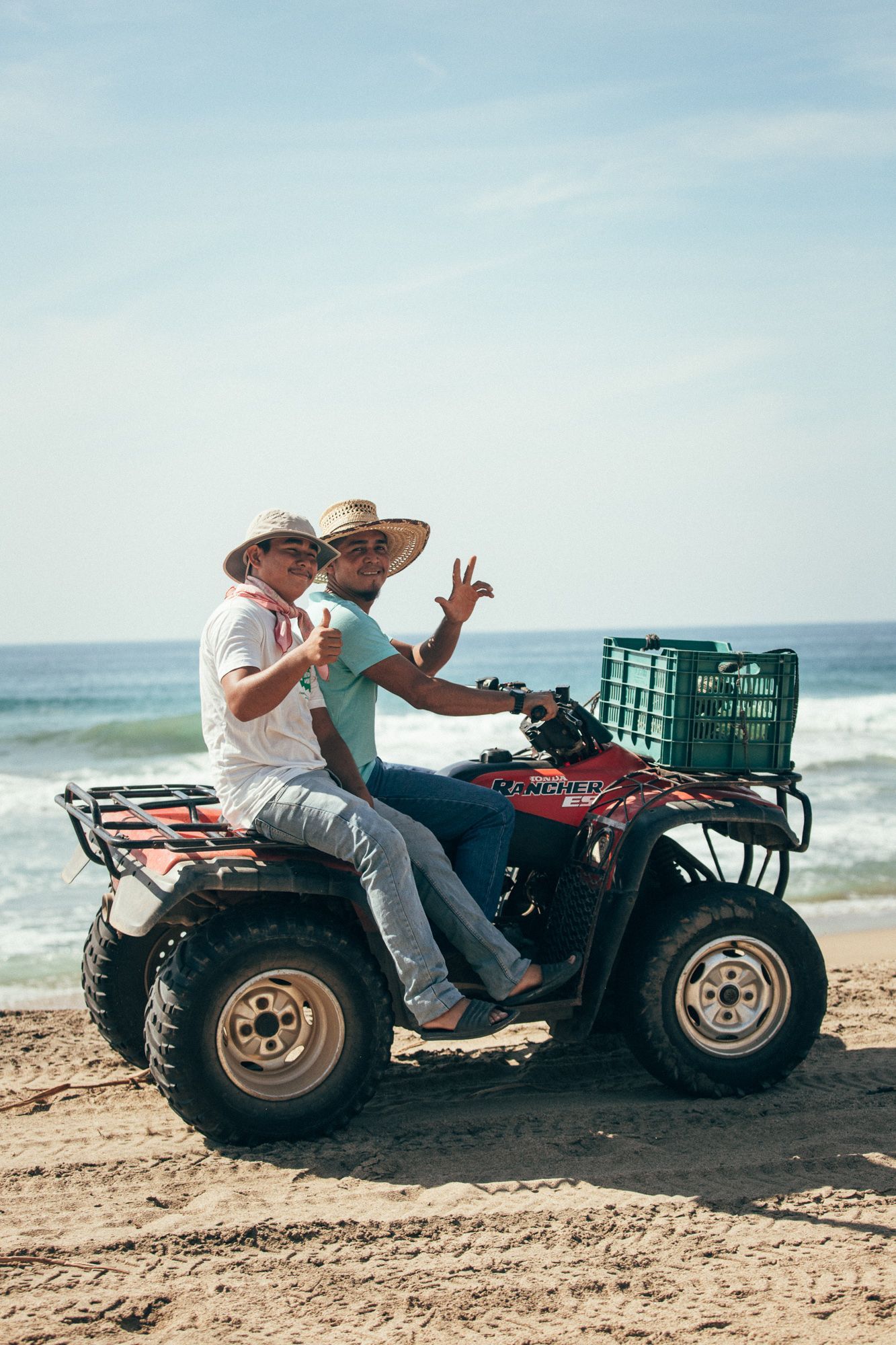 Two people riding an ATV on a beach, waving near the ocean with a crate on the back.