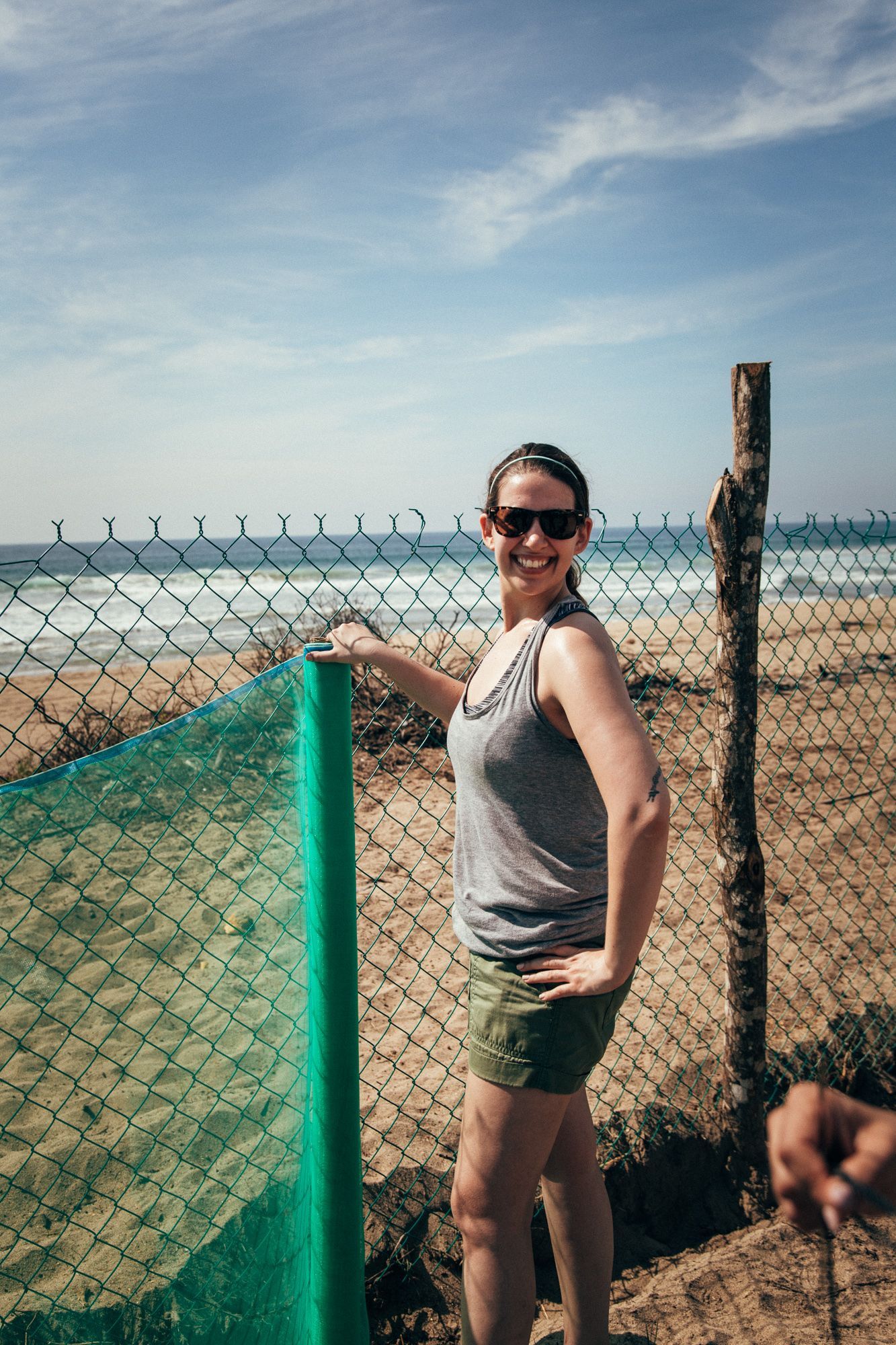 Person smiling by a chain-link fence at a beach, with sand, sea, and blue sky in the background.