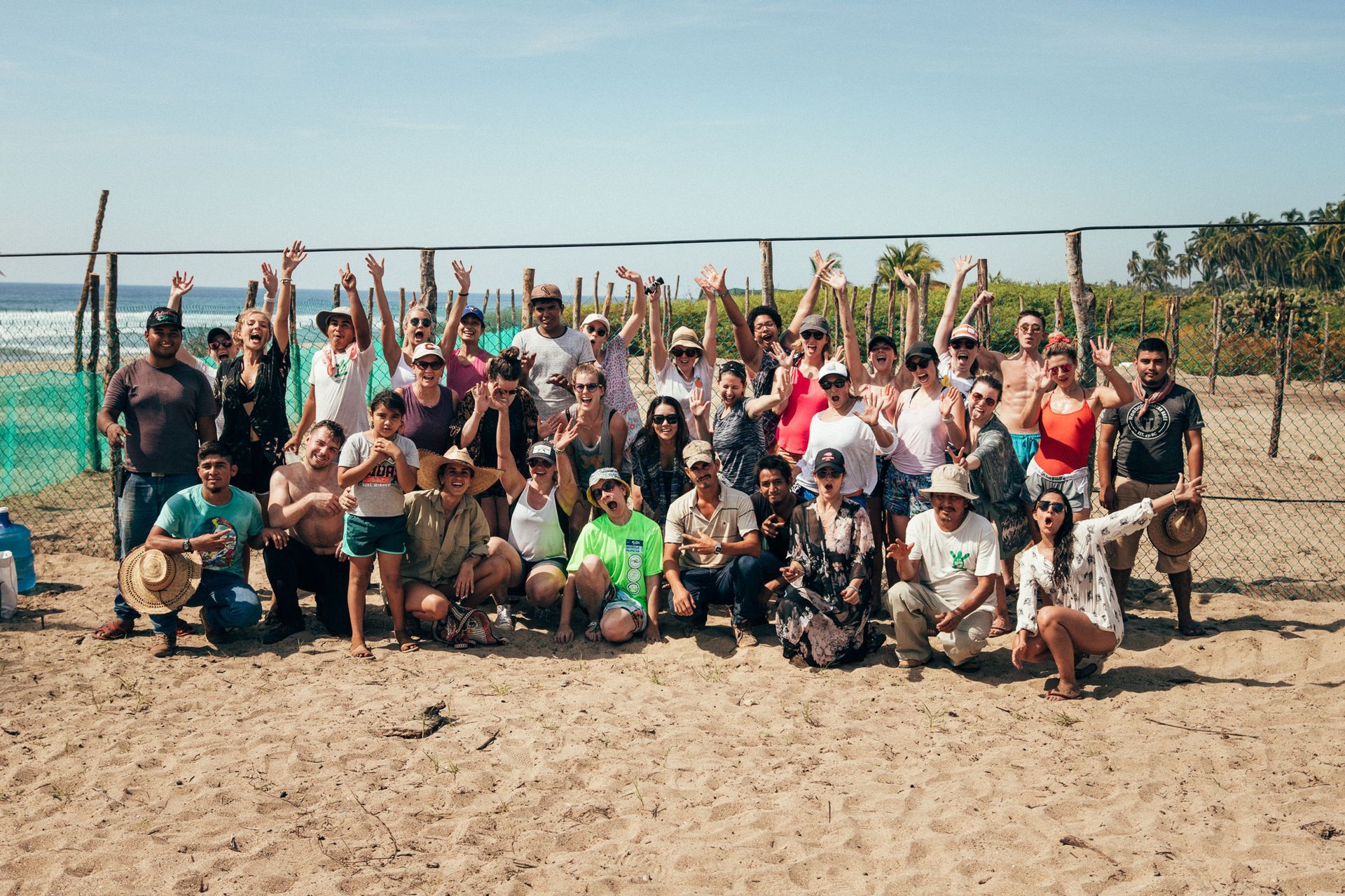 Group posing and cheering on a sandy beach with volleyball net and ocean behind them
