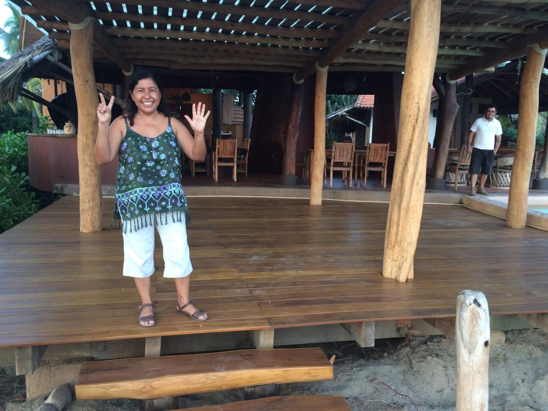 Smiling woman waving on a wooden deck under a thatched roof, with chairs in the background.