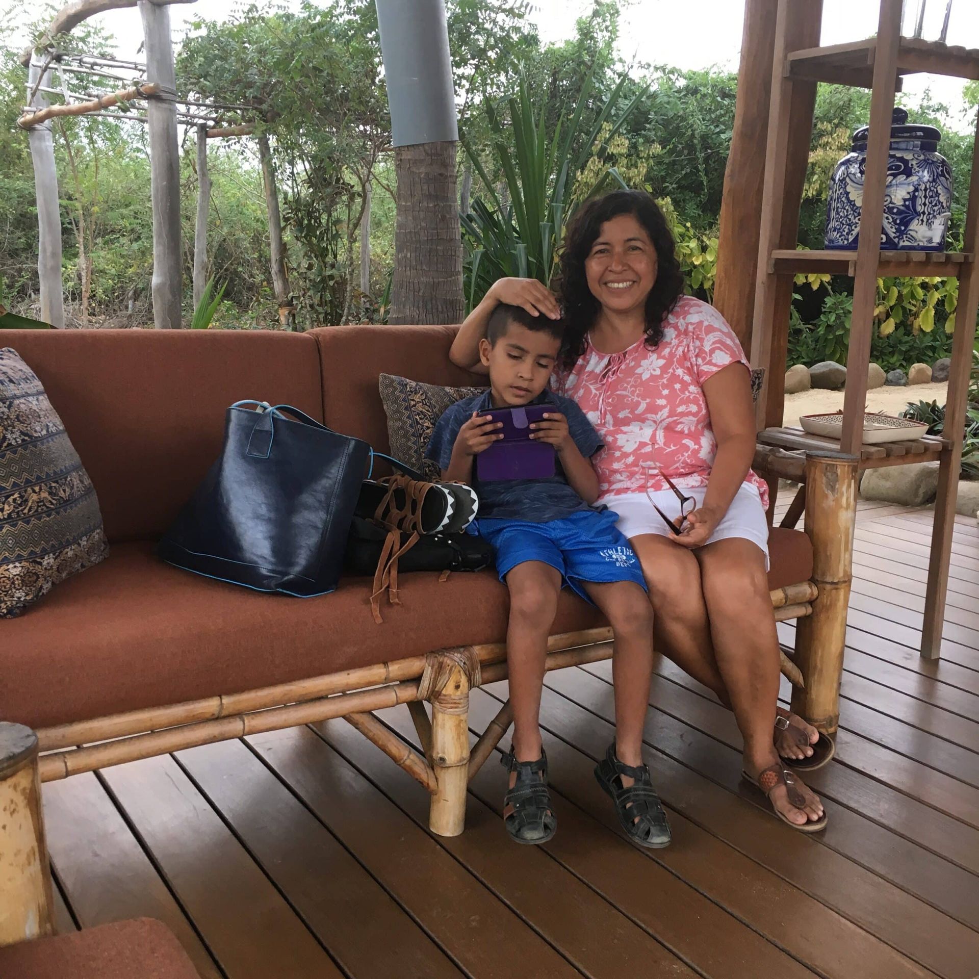 Woman and child sitting on a wicker bench on a wooden patio, smiling with a lush garden in the background