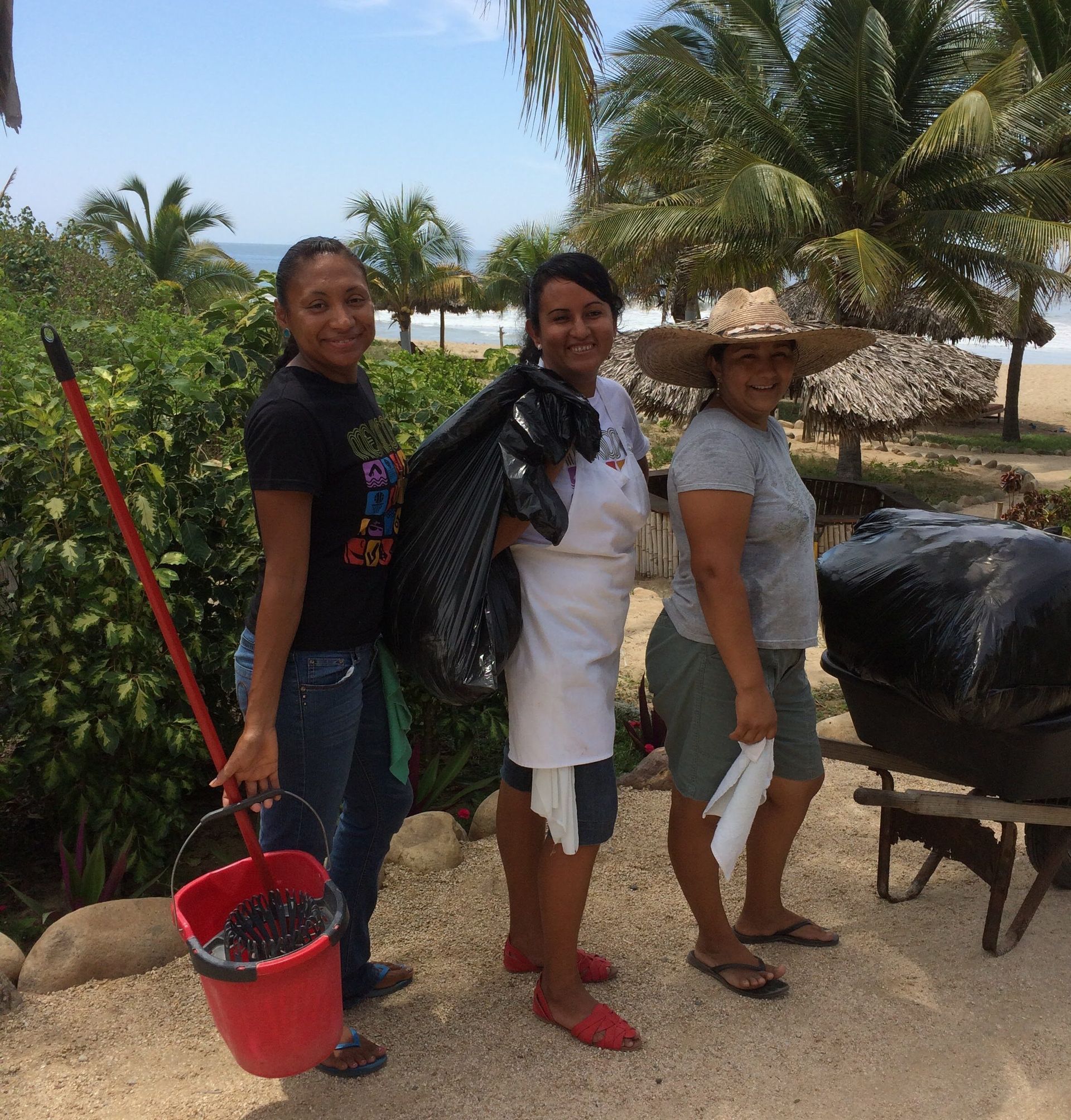 Three people stand by a beach cart under palm trees, one holding a red mop bucket.