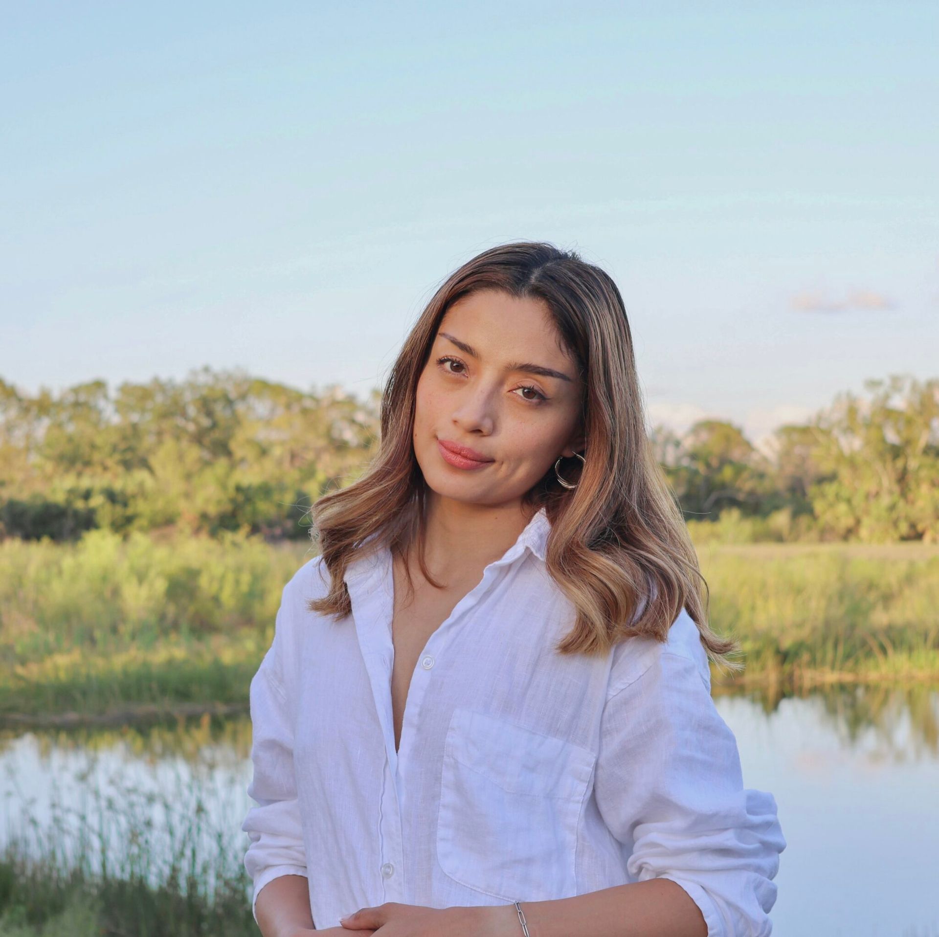Woman in a white shirt standing by a calm lake with trees and a clear sky in the background