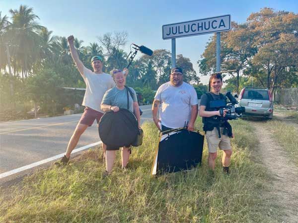 Four people posing by a roadside sign reading “JULUCUCA” in a sunny rural setting