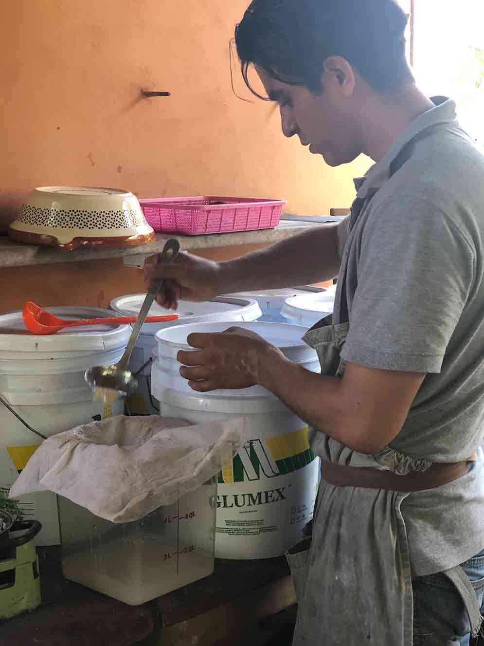 Person stirring a white bucket in a kitchen, with cooking containers and utensils on a counter.