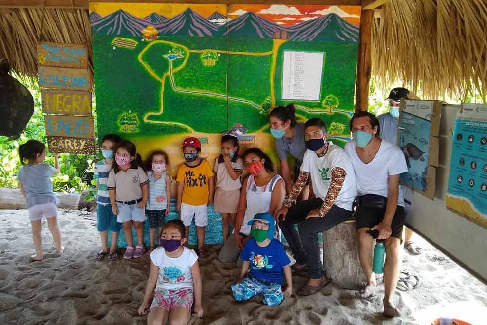 Children and adults posing under a thatched shelter with a colorful map mural in the background
