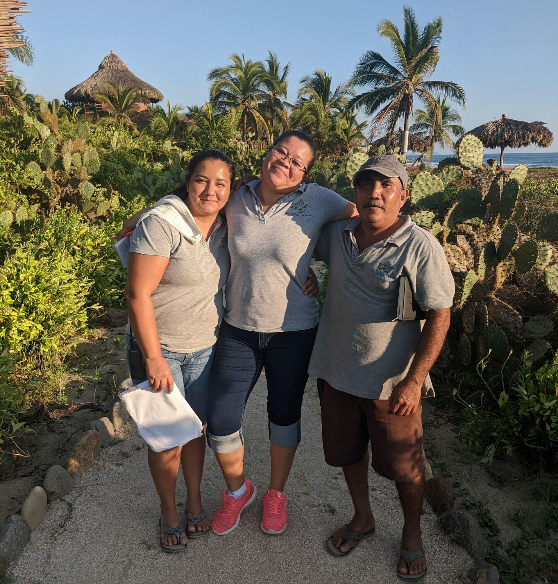 Three people posing on a sunny path with tropical plants and palm trees in the background.