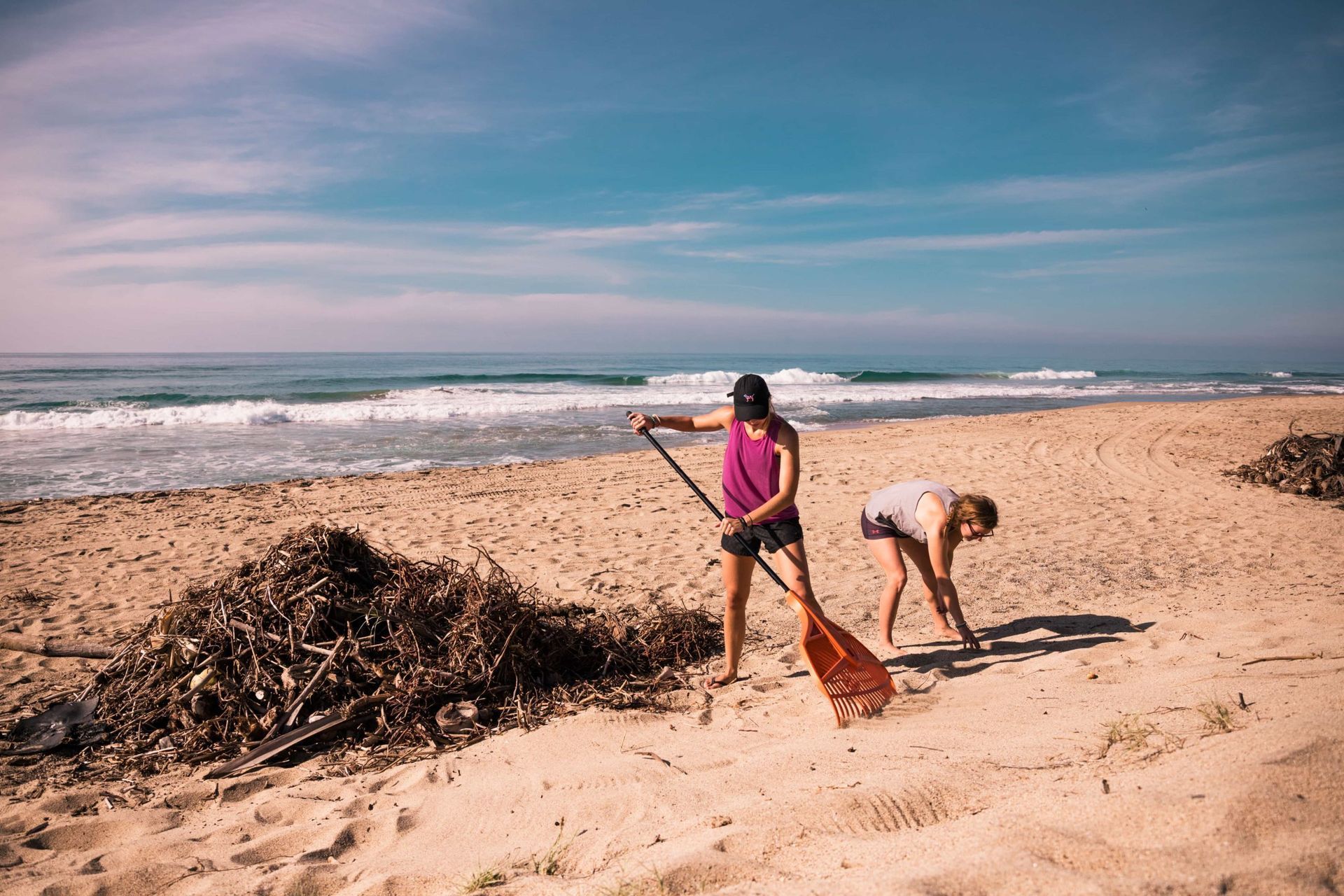 Two people clean debris on a sandy beach beside the ocean.