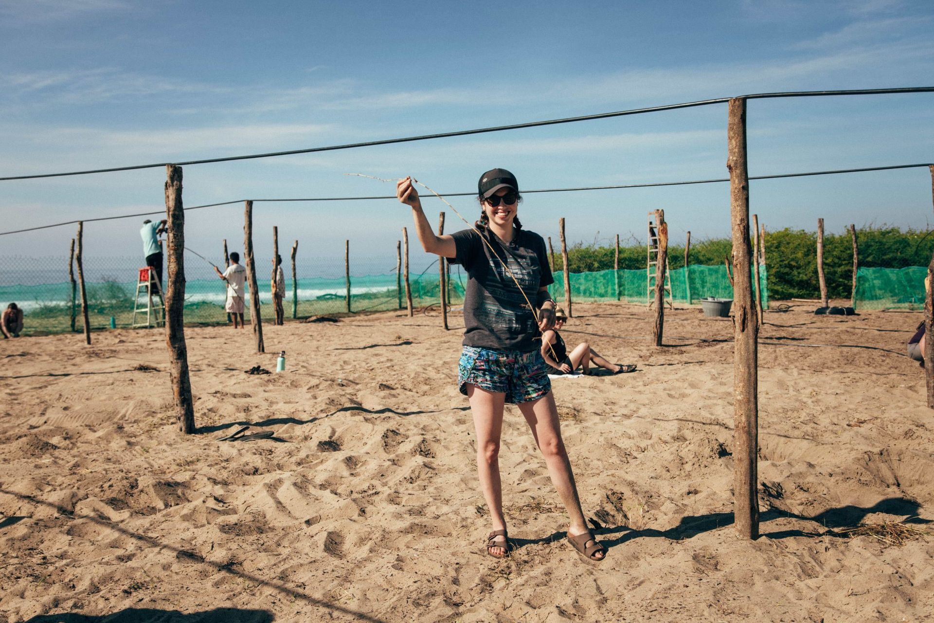 Person standing in a sandy outdoor obstacle course, holding a rope between wooden posts under a clear sky