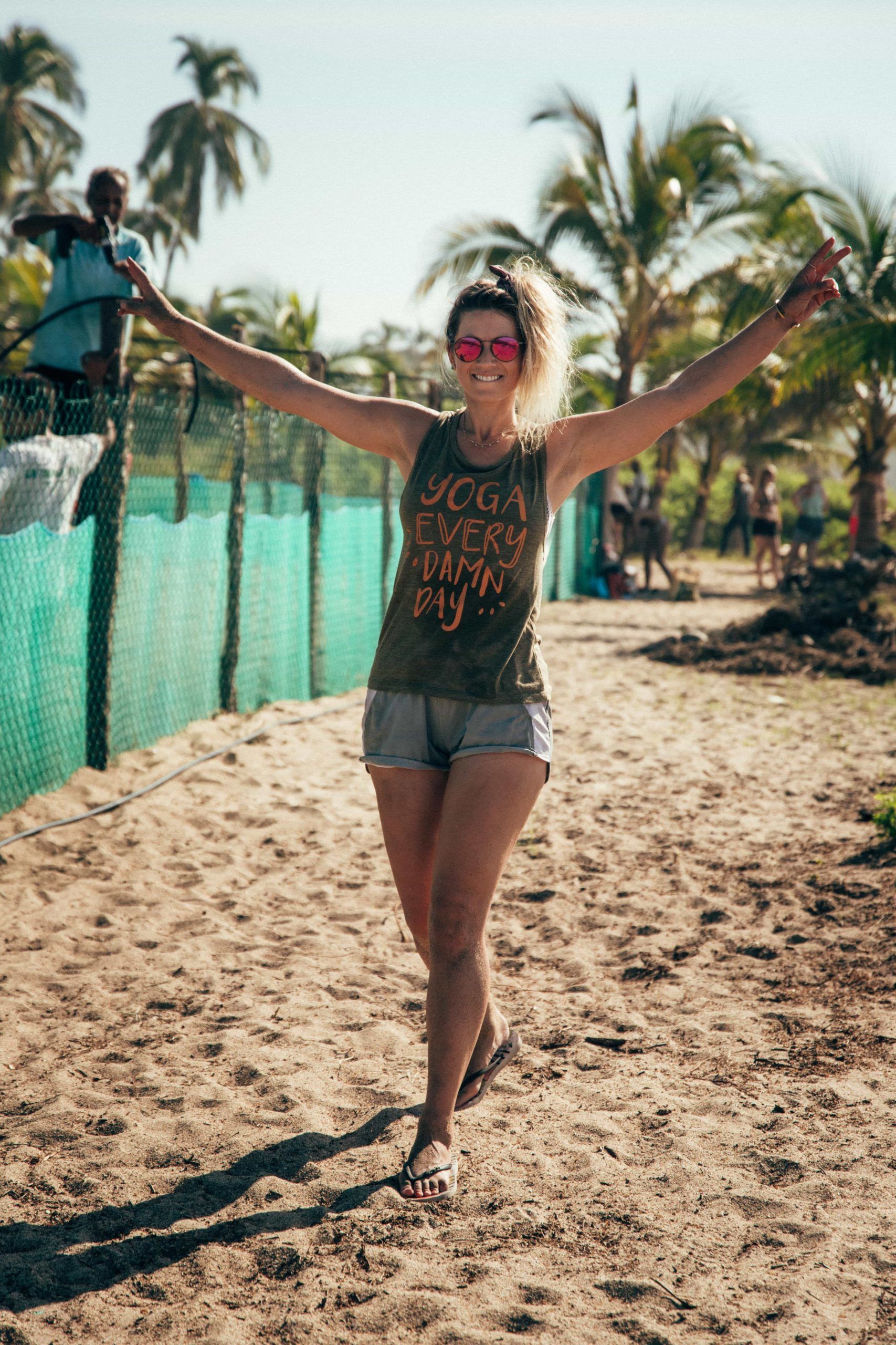 Woman walking on a sandy beach with arms raised, palm trees and turquoise water behind her.
