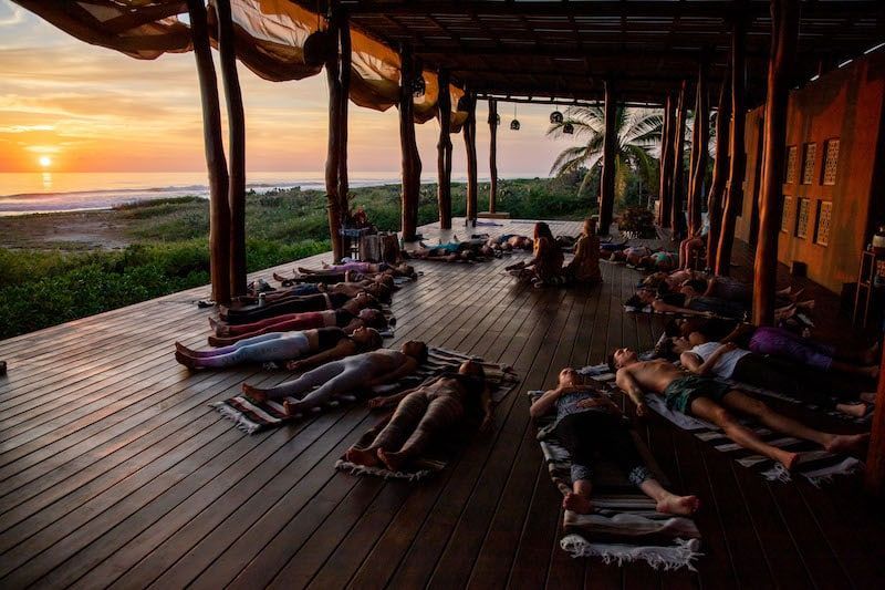 People lying on yoga mats in an open-air wooden pavilion at sunset, overlooking a beach.