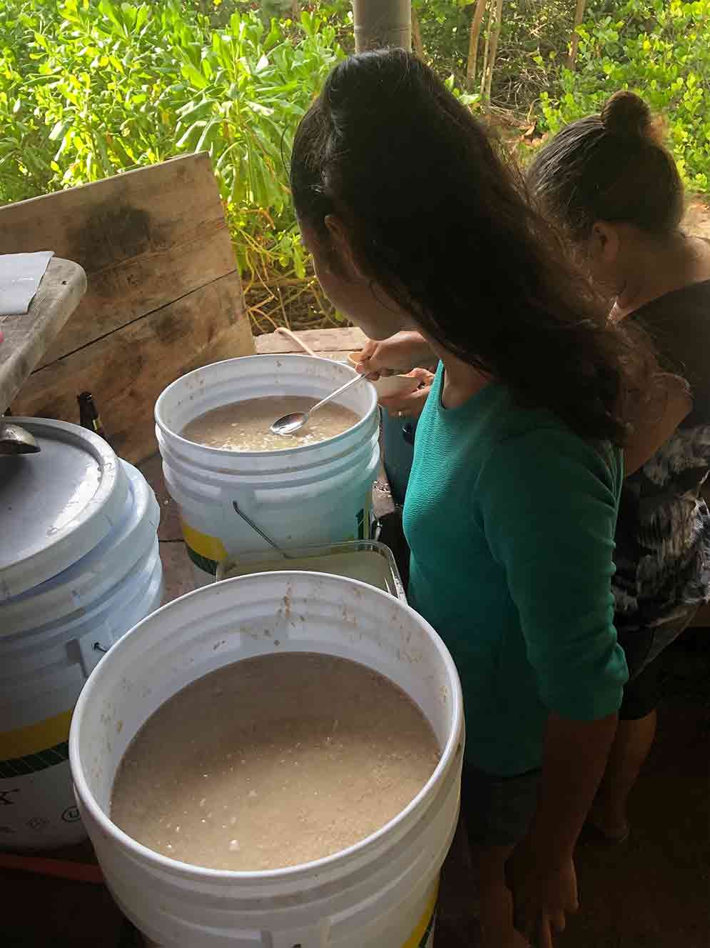 Two children near large white buckets of beige liquid in a rustic outdoor setting