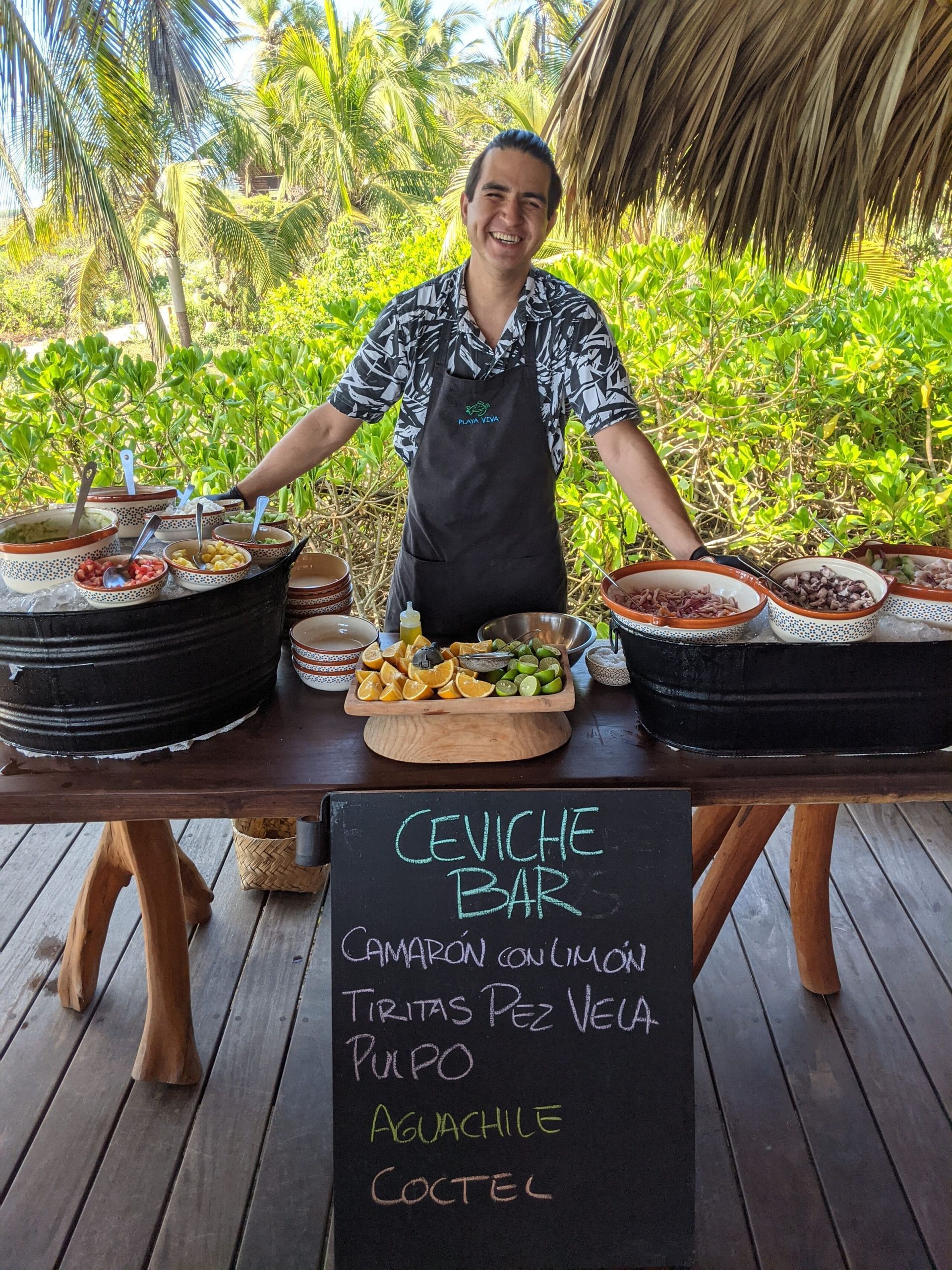 Person smiling behind a food stall with trays of food and a chalkboard menu outdoors under a thatched roof