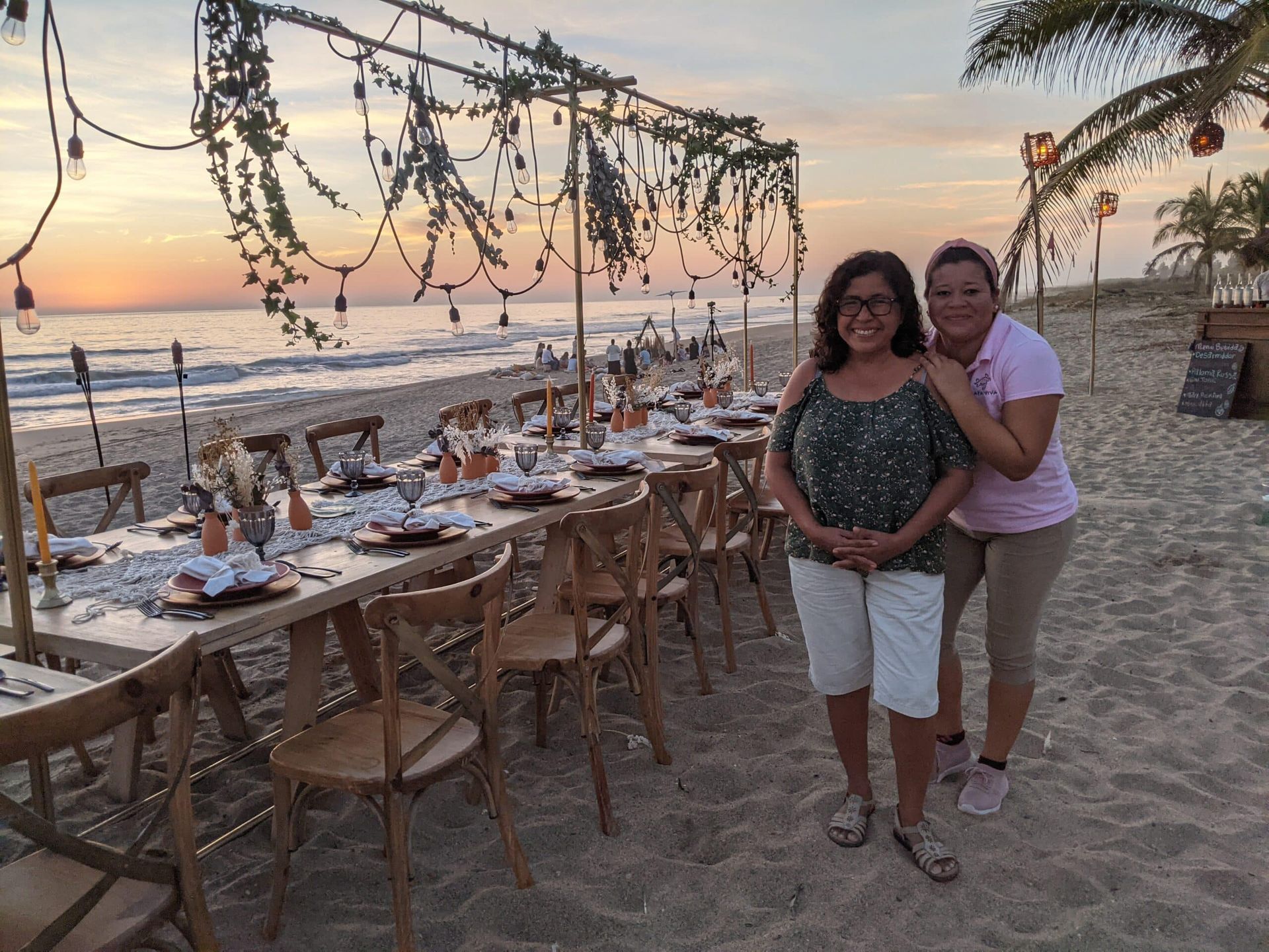 Two people standing on a decorated beach dinner setup at sunset with the ocean in the background