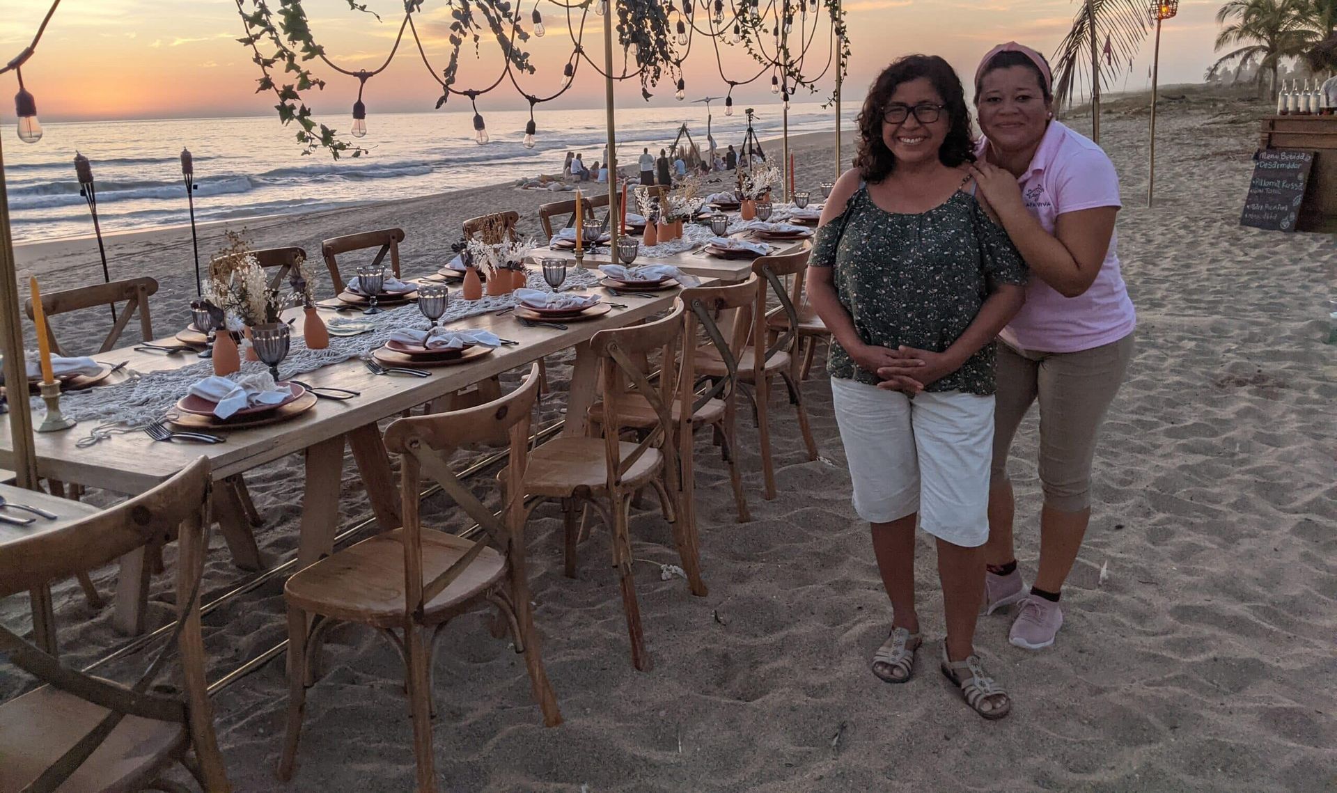 Two women standing beside a beachside dinner table setup at sunset with ocean view and string lights