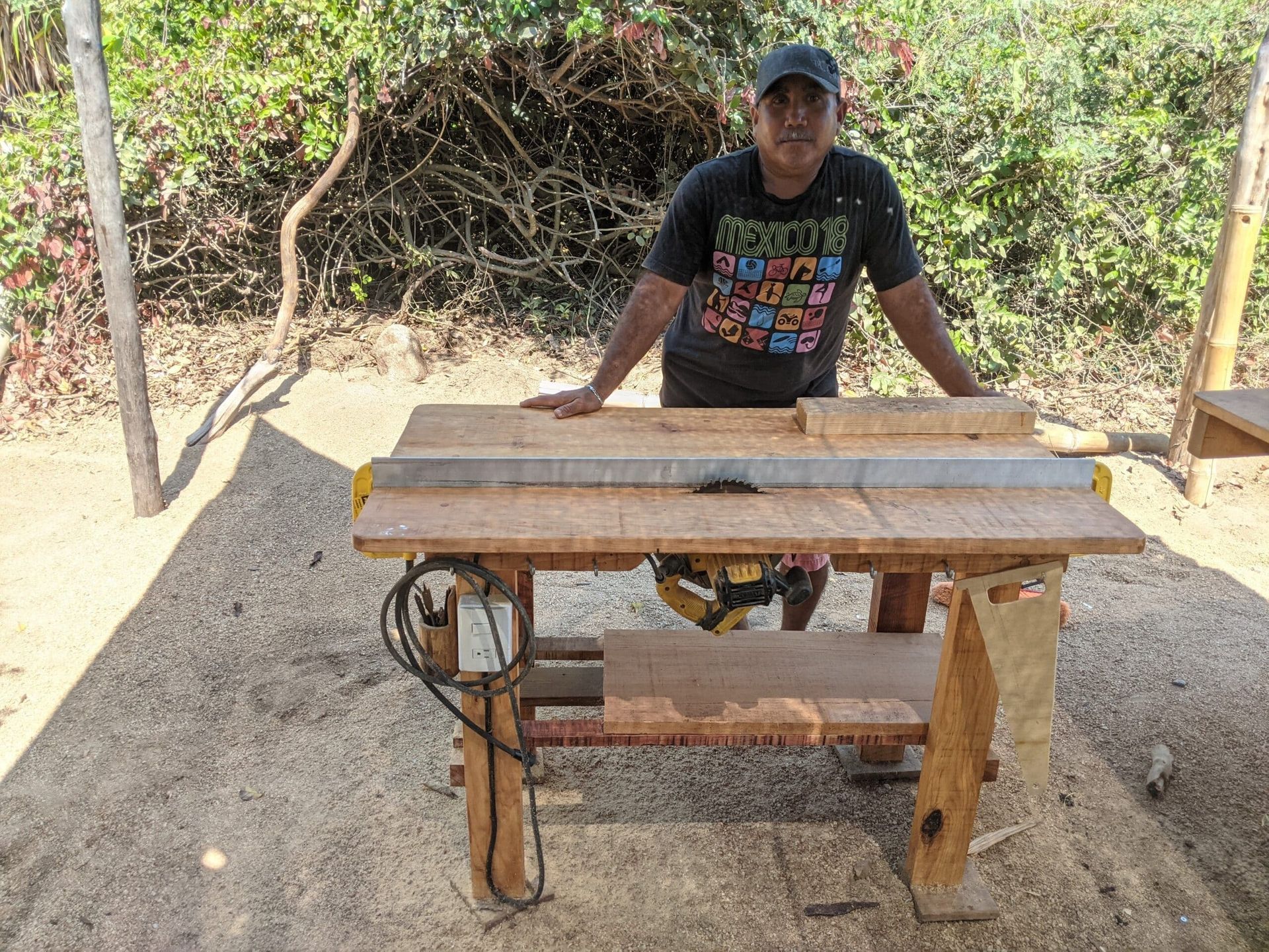 Man standing behind a wooden workbench outdoors, in front of dry vegetation and dirt ground.