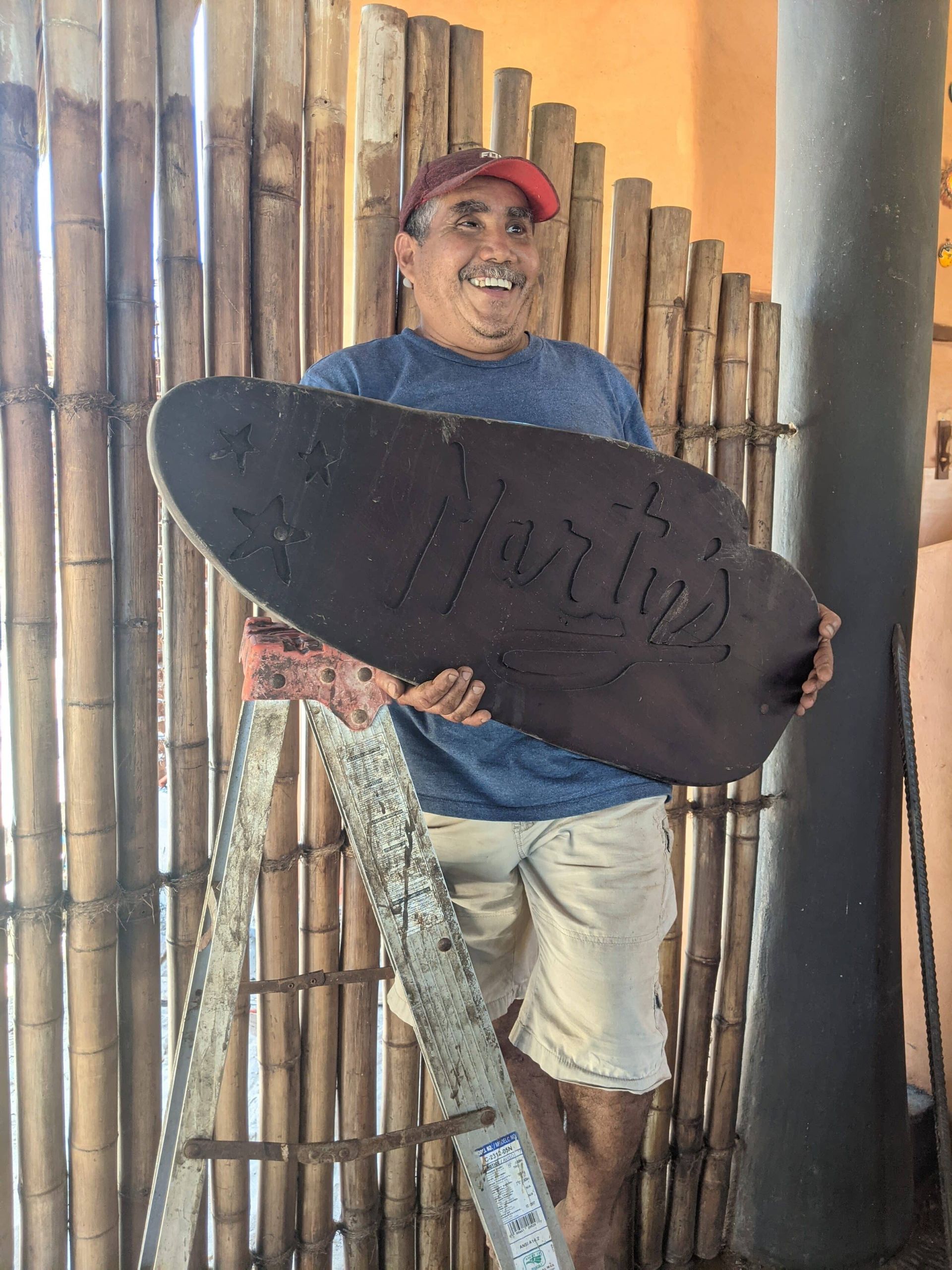 Smiling person holding a large black surfboard-shaped sign beside bamboo fencing
